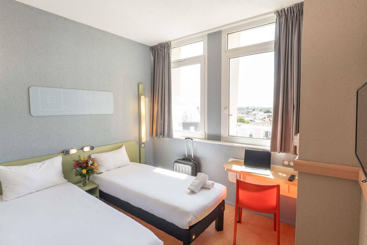 A hotel room with two beds, a desk with a laptop, a chair, a window with curtains, and a wall-mounted light fixture.
