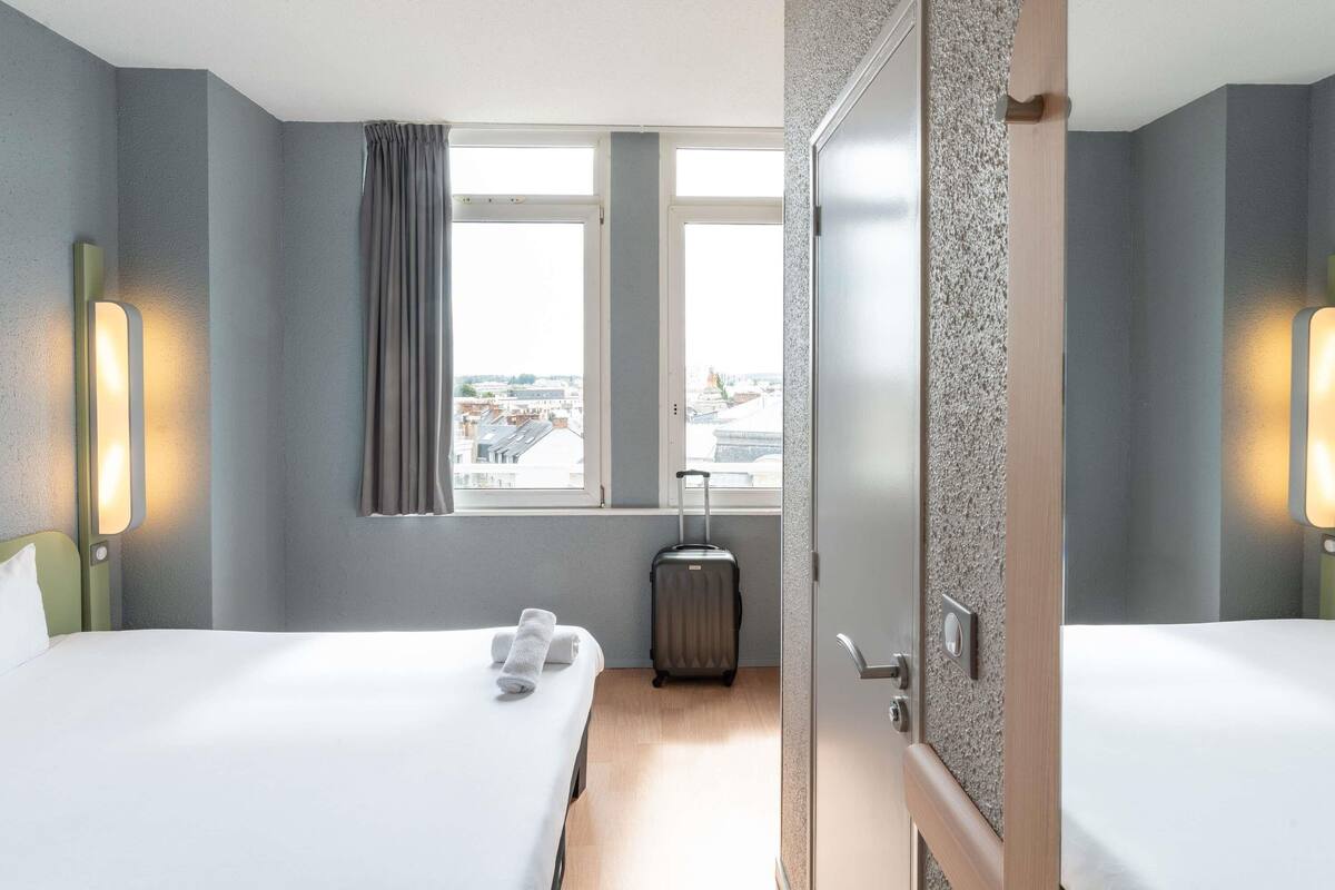 A hotel room with two beds, a window with curtains, a suitcase, and a door.