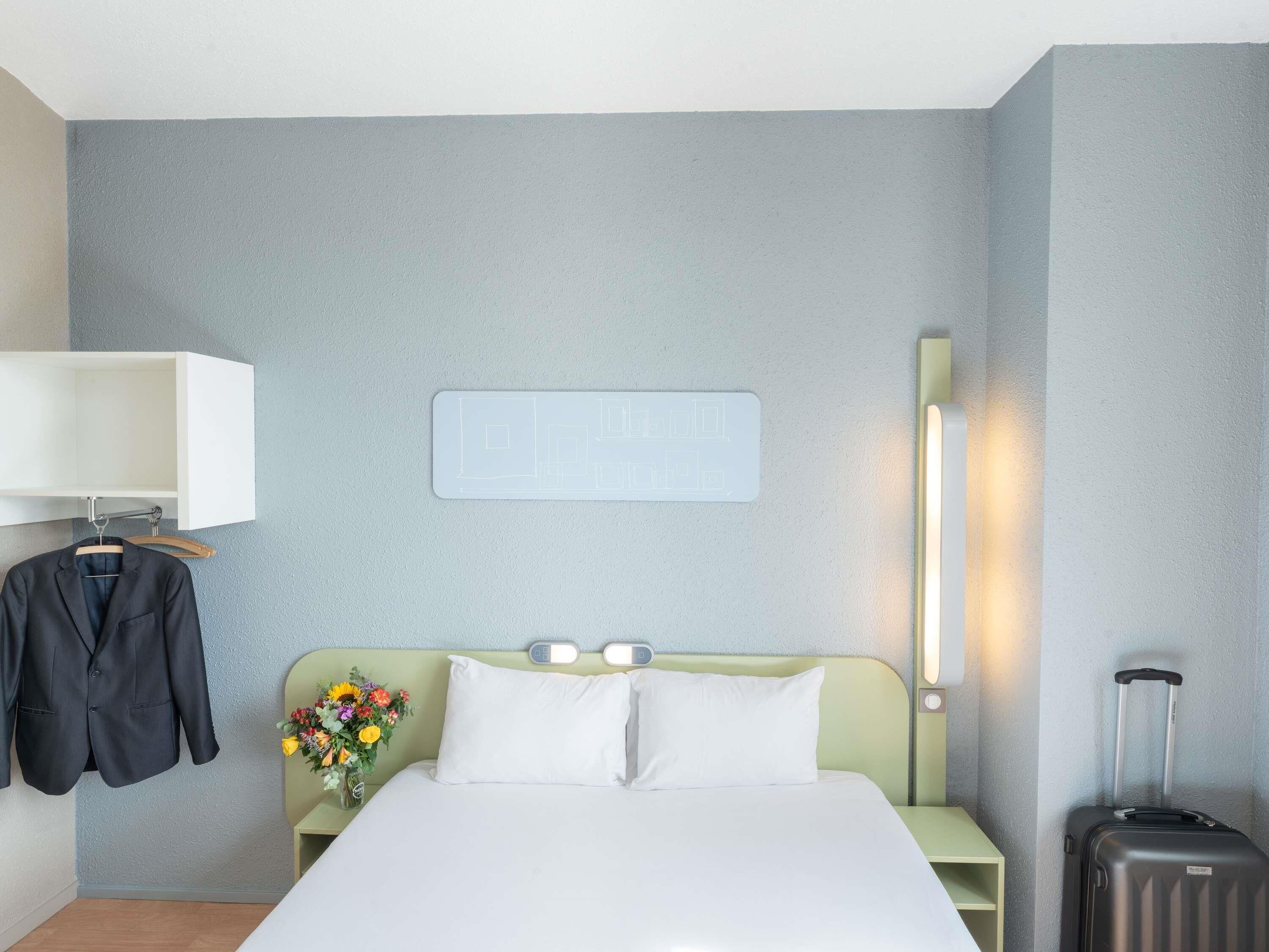 Habitación de hotel con cama, mesita de noche con un jarrón de flores, perchero y lámpara de pared, además de una maleta.