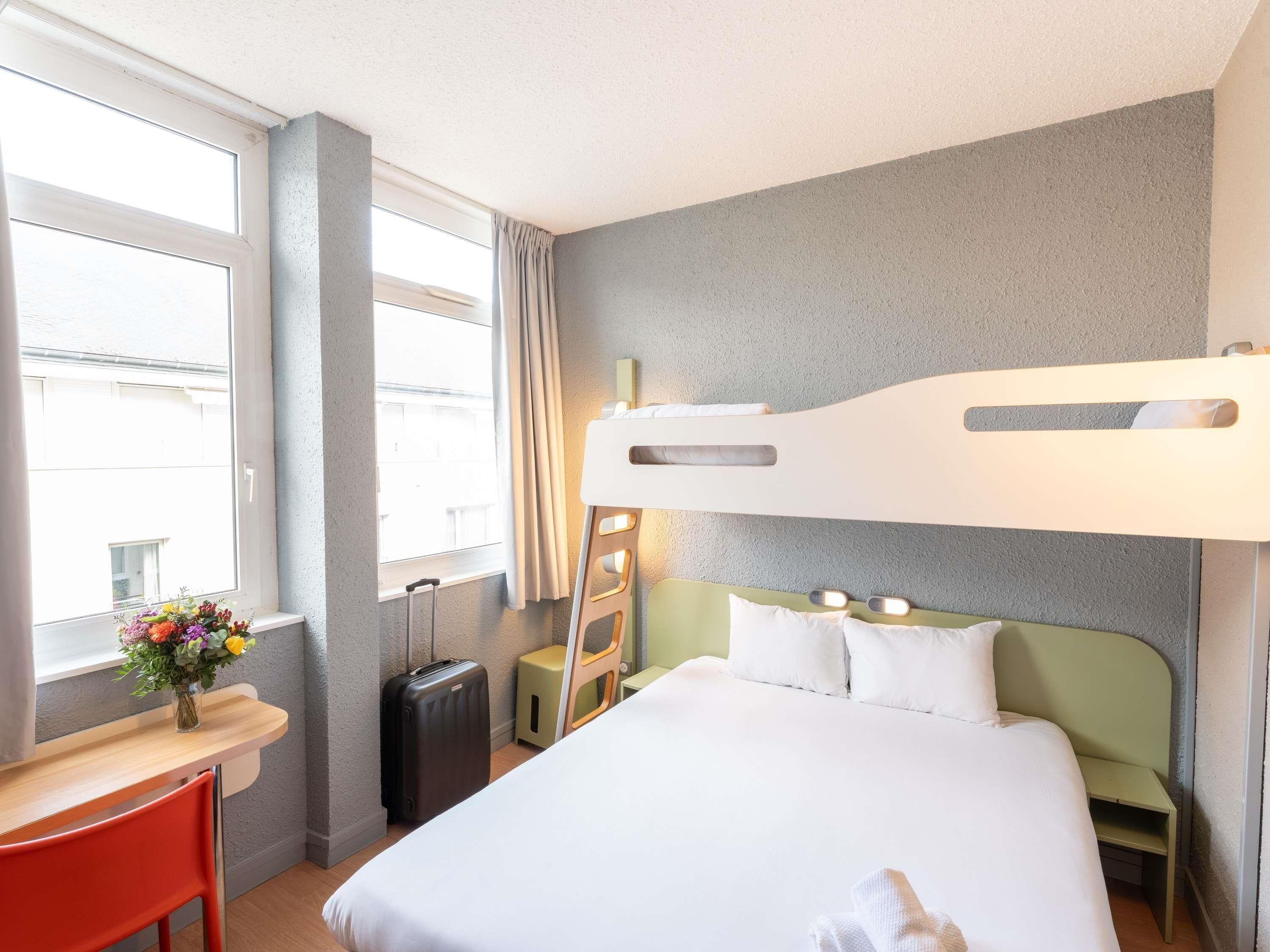 Une chambre d’hôtel avec un lit superposé, un bureau avec une chaise, une valise et un vase de fleurs placé près de la fenêtre.