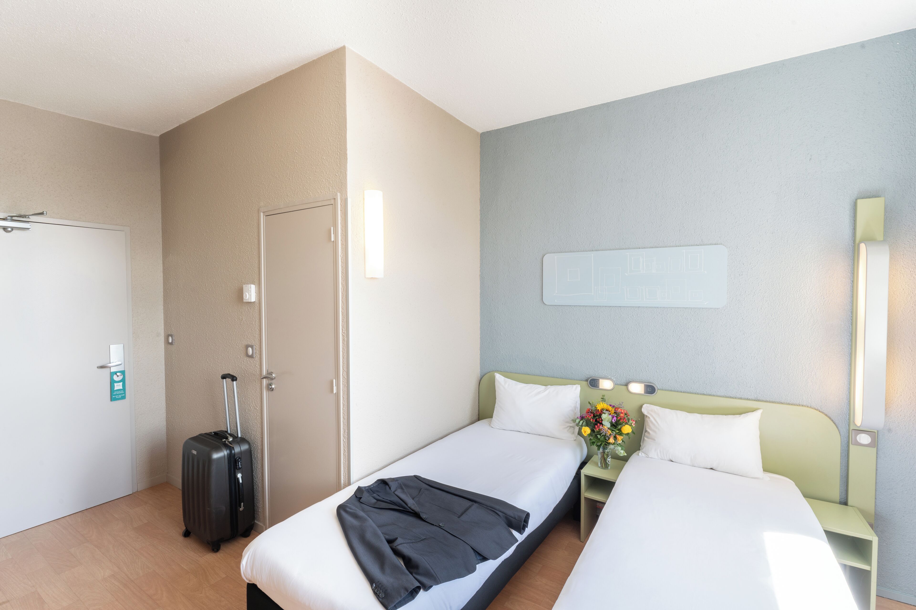 A hotel room with two single beds, a small bedside table with a vase of flowers, a wall-mounted light, and a suitcase on the floor.