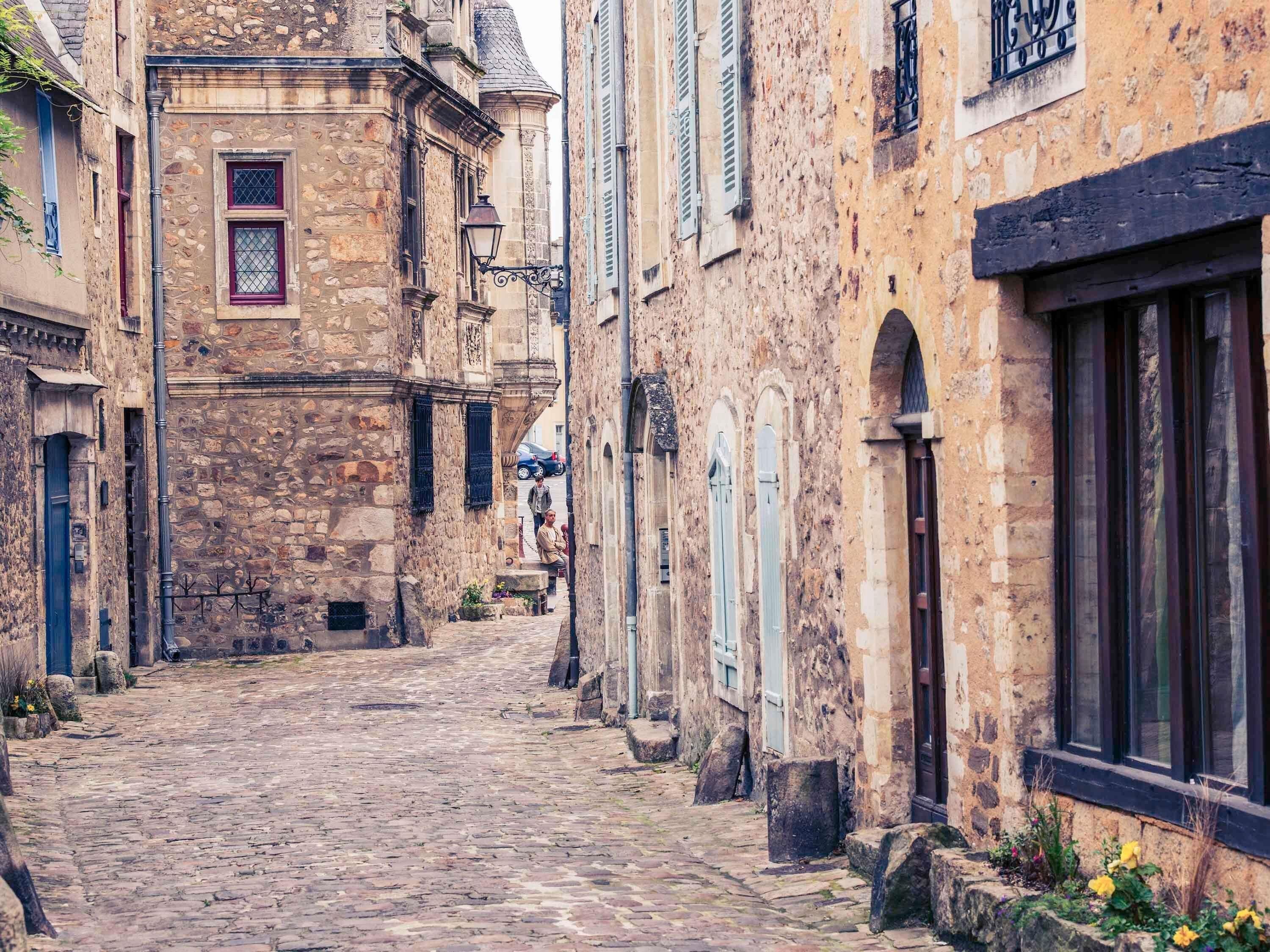 A narrow cobblestone street flanked by historic stone buildings with arched windows and shutters.