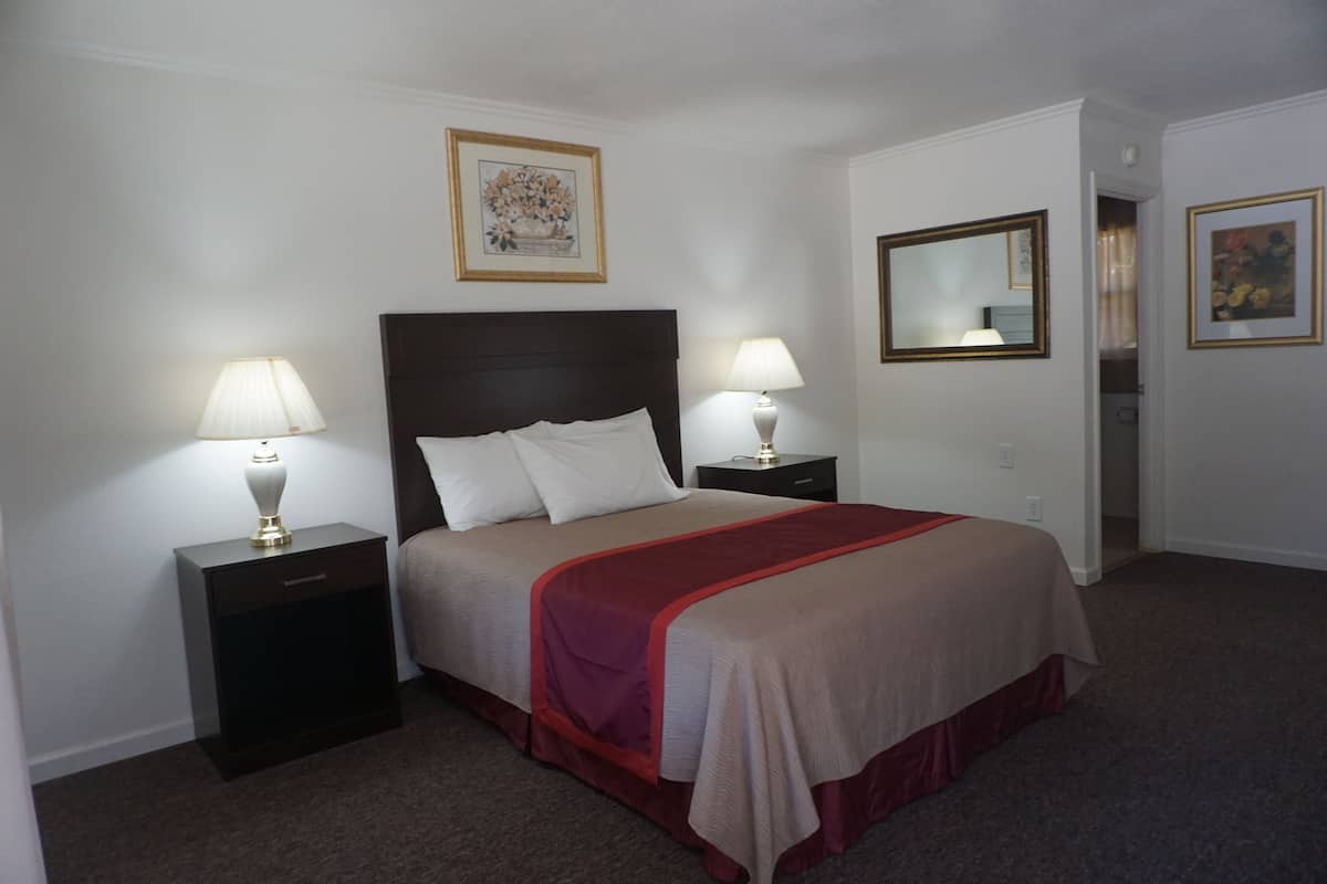 A neatly made bed with a red and beige bedspread, two bedside lamps, a framed floral artwork above the bed, and a mirror on the wall.