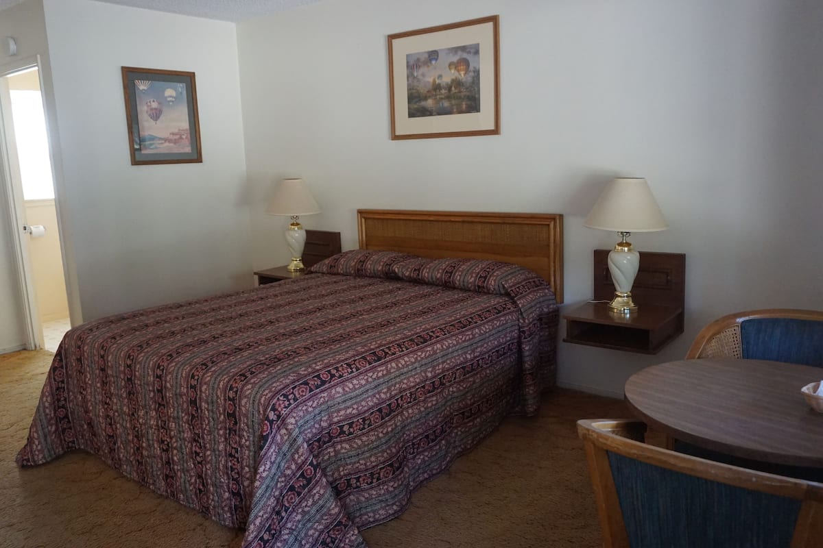 A hotel room with a large bed, two bedside tables with lamps, a small round table with a chair, and two framed pictures on the walls.