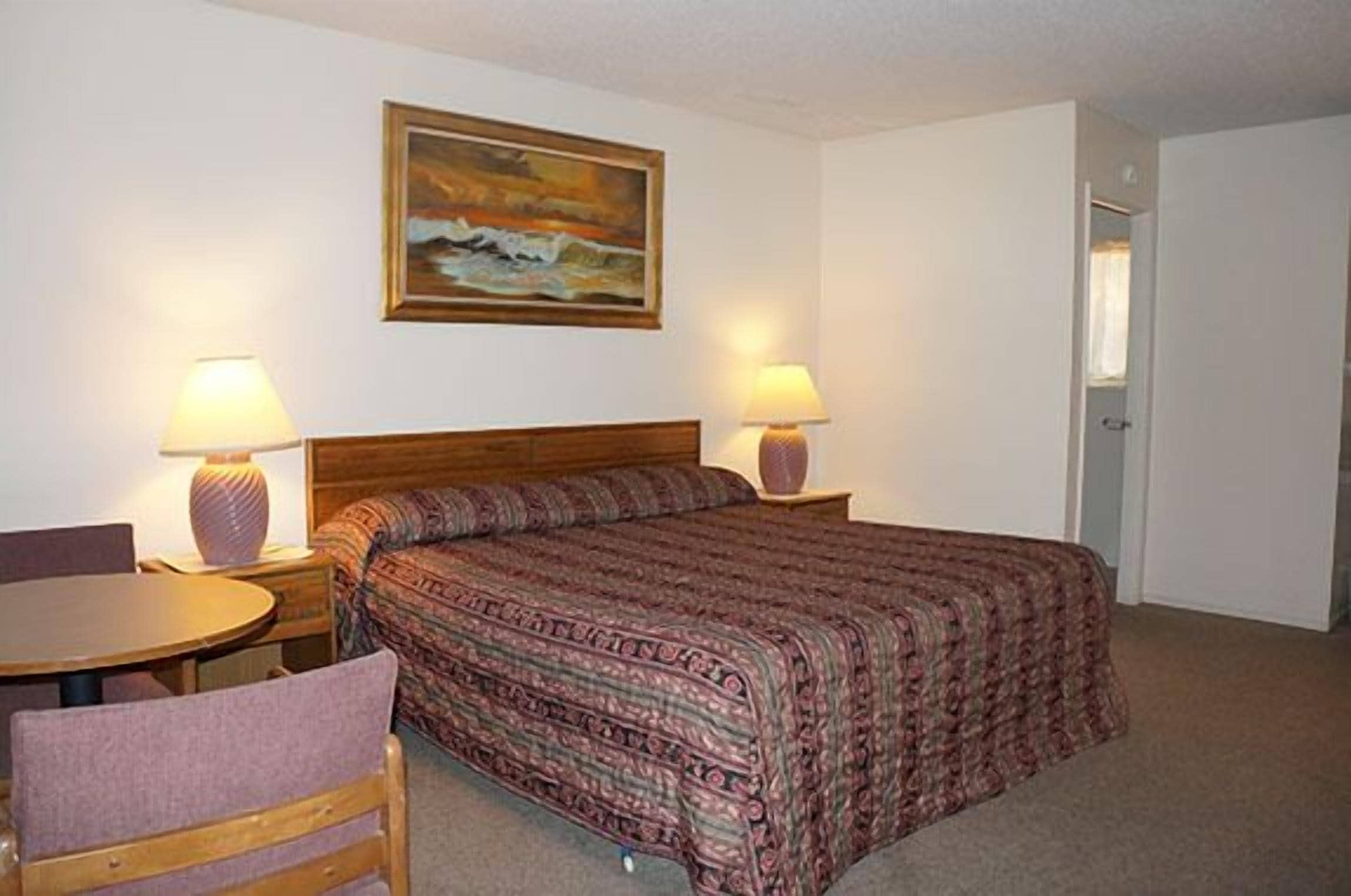 A hotel room with a bed, two bedside lamps, a painting on the wall, and a small table with a chair.