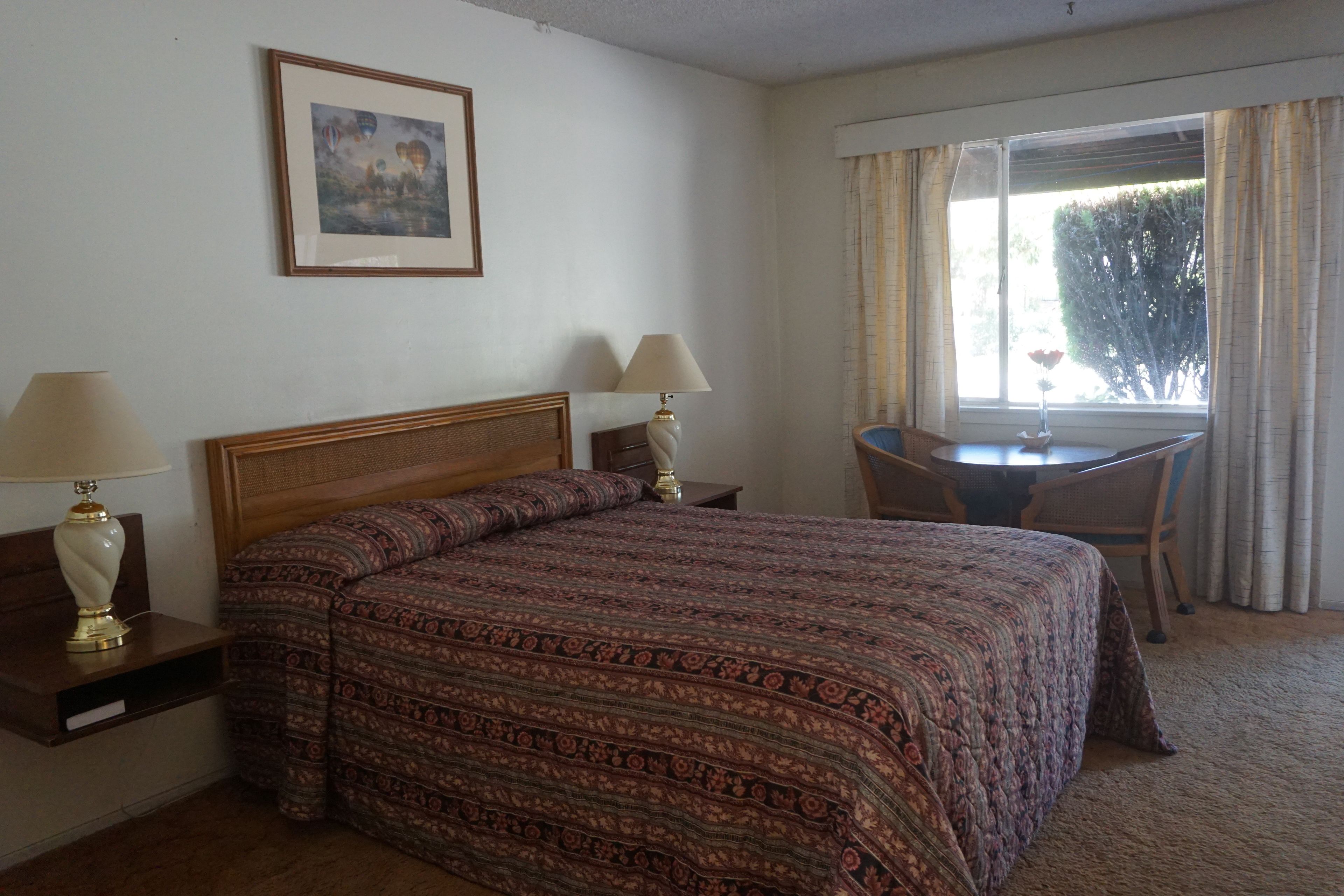 Ein Hotelzimmer mit einem Bett, zwei Nachttischlampen, einem gerahmten Bild an der Wand, einem Esstisch mit Stühlen und einem Fenster mit Vorhängen.