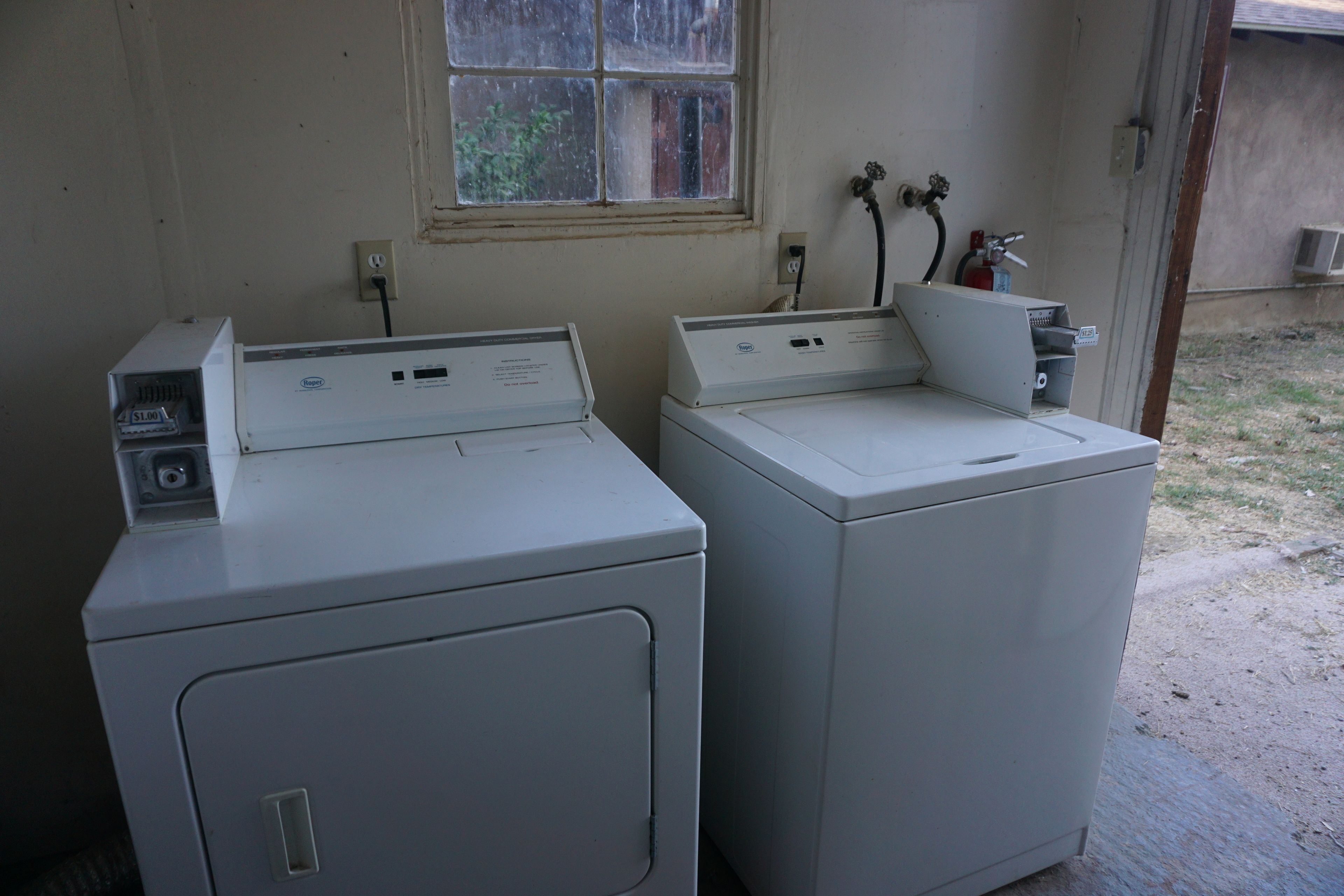 Two white front-loading washing machines in a laundry room with a window, electrical outlets, and a hose.