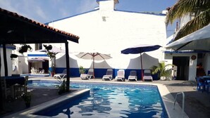 A swimming pool area with lounge chairs and umbrellas, surrounded by a white building with blue trim. - Hotel y Bungalows Los Arcos (San Patricio Melaque)