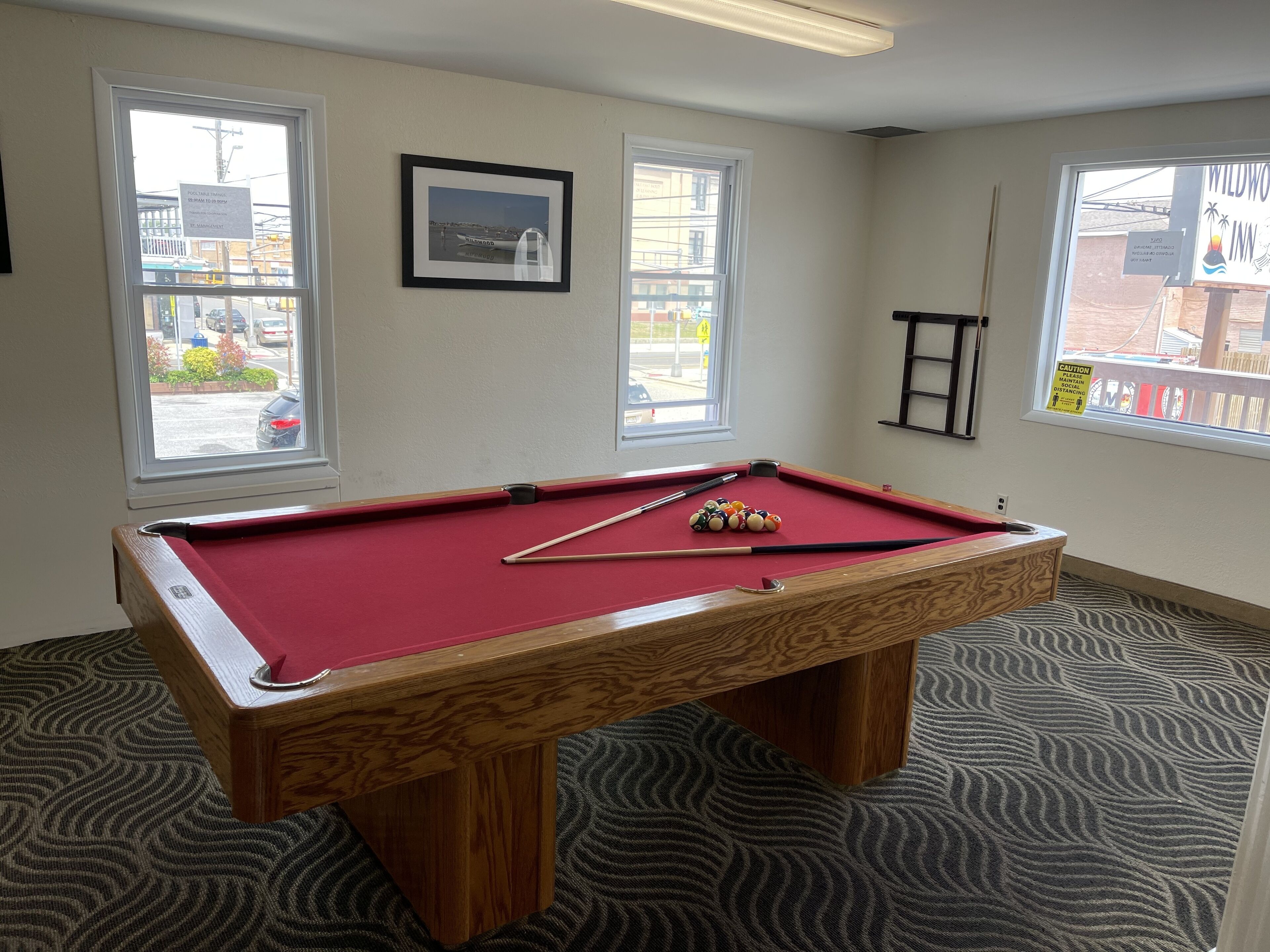 A pool table with balls and cues, a framed picture on the wall, and windows with views of the outside.