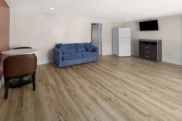 A modern, minimalistic room with a wooden floor, a blue sofa, a small round table, a white refrigerator, and a TV mounted on the wall.