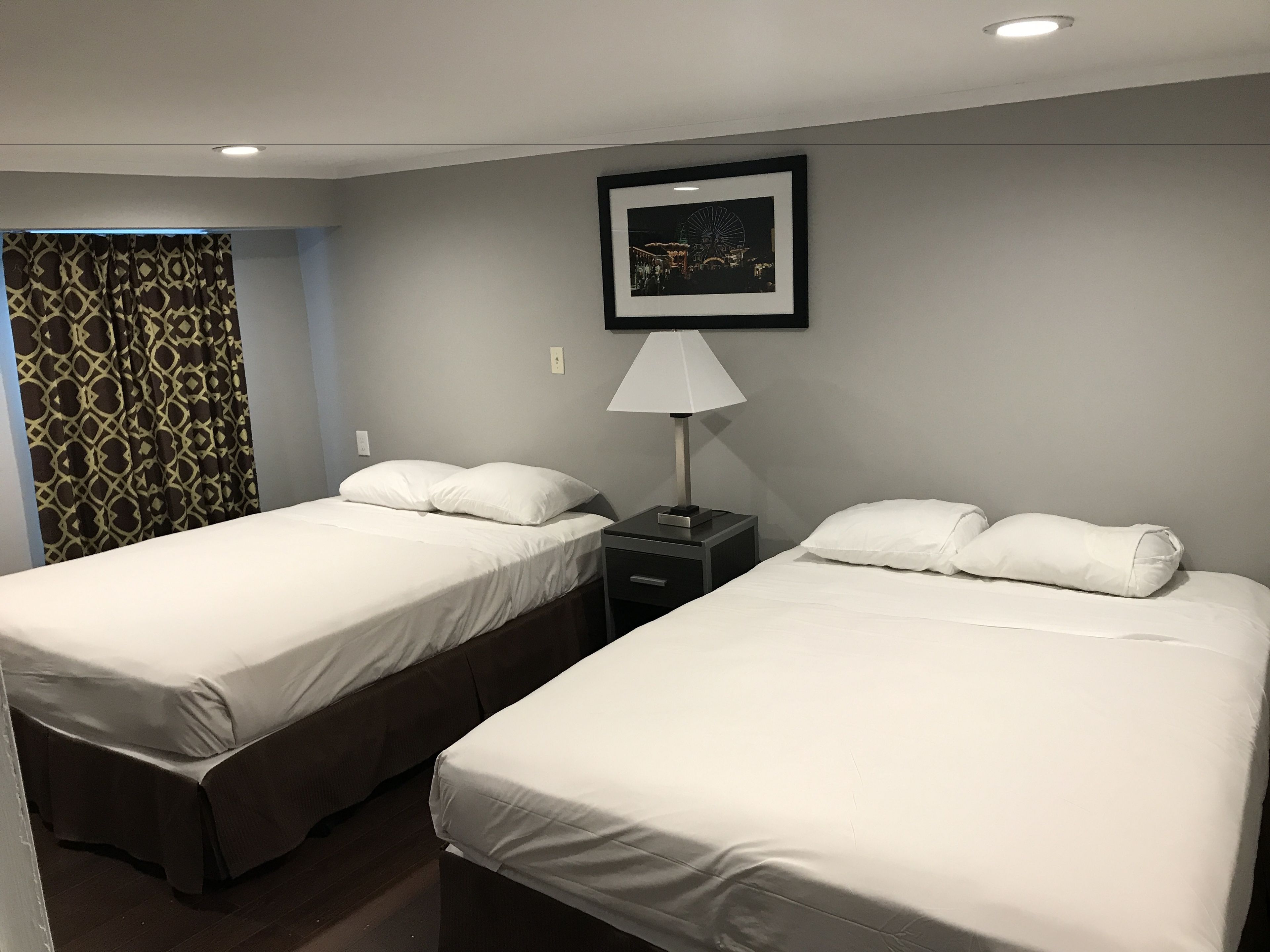 Een hotelkamer met twee bedden, een nachtkastje met een lamp, een lijst met een foto aan de muur, en kamergordijnen met een patroon.