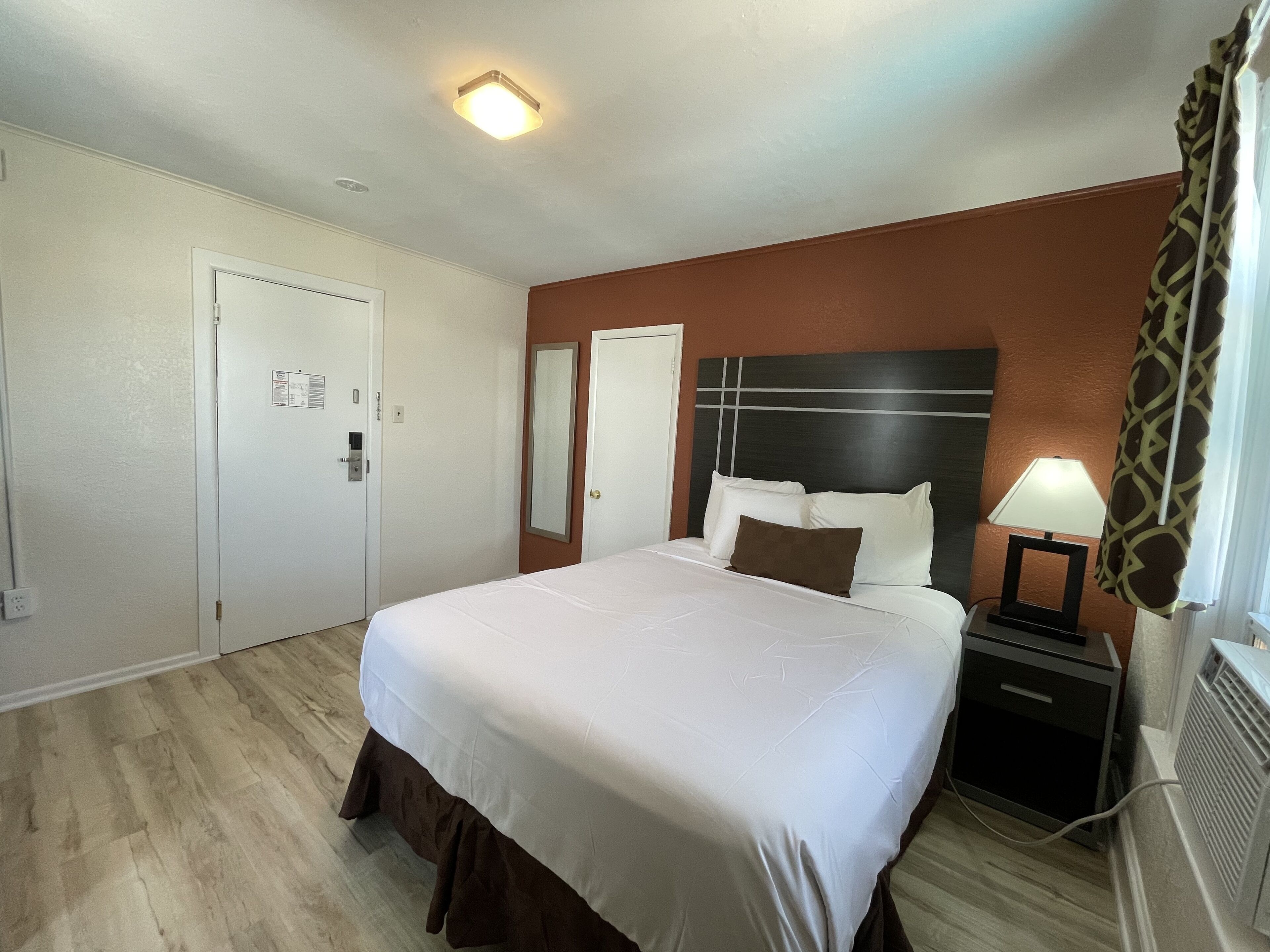 A hotel room with a large bed, a nightstand with a lamp, a mirror, and a door.