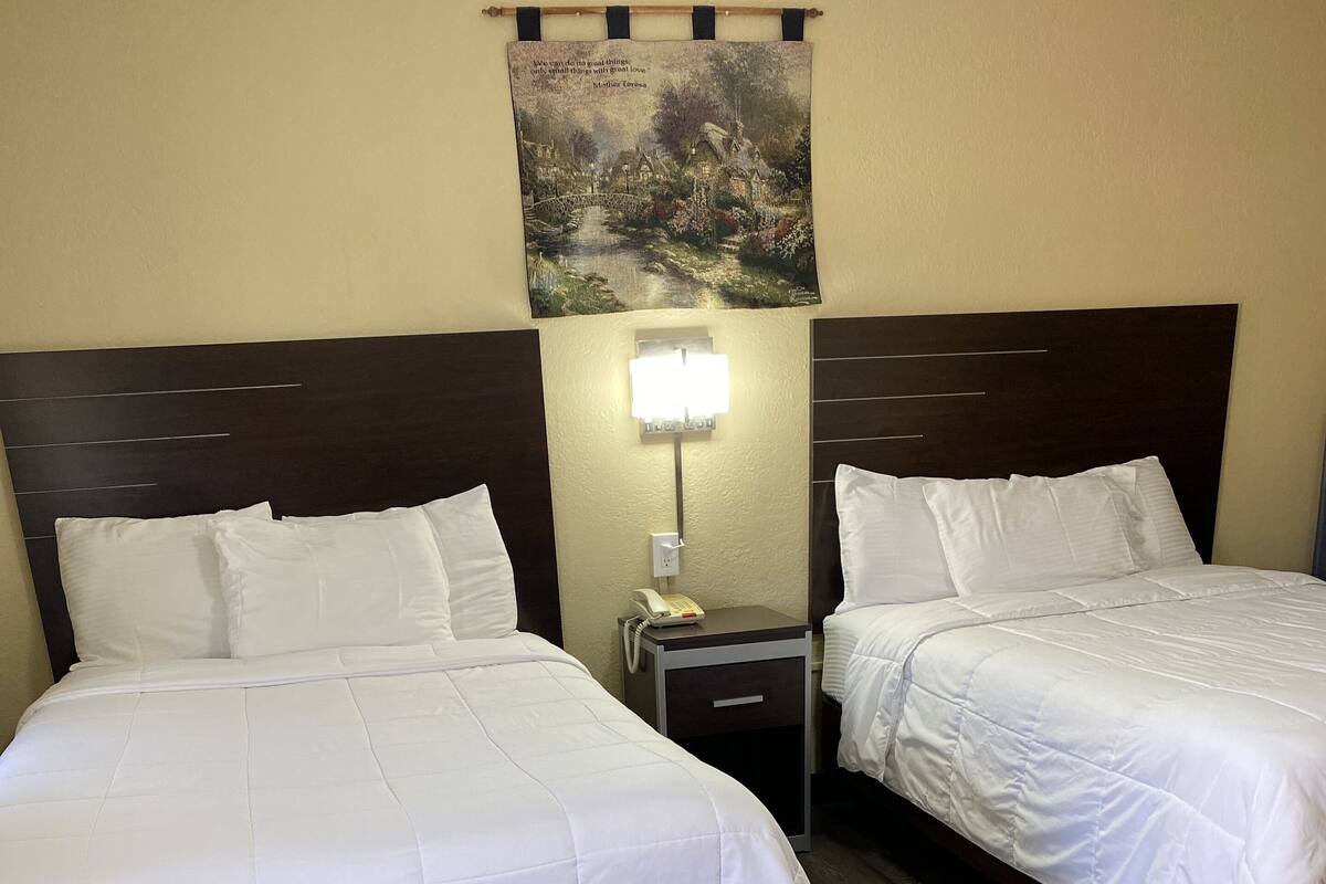 A hotel room with two beds, a bedside table with a lamp, and a wall-mounted painting.
