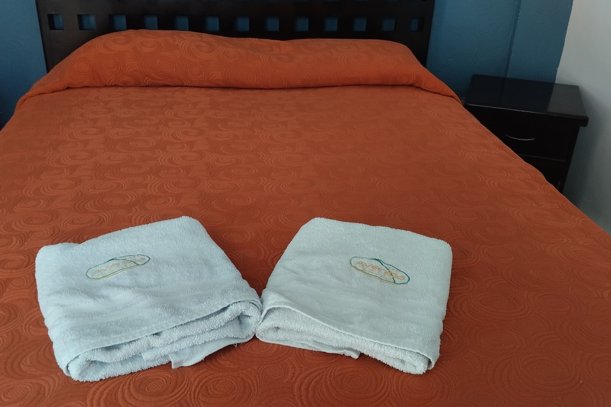 A bed with an orange bedspread, two folded towels with a logo, a black nightstand, and a blue wall.