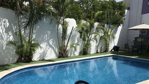 Una piscina con bordo curvo, circondata da erba sintetica verde e palme. In primo piano, una sedia in metallo nero.
