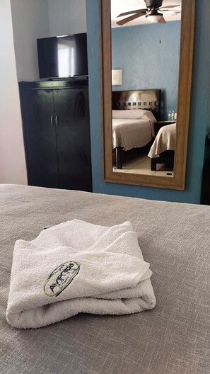 A neatly folded towel with a logo on a bed in a room with a dark wardrobe and a ceiling fan. - Hotel Avenida (Campeche)