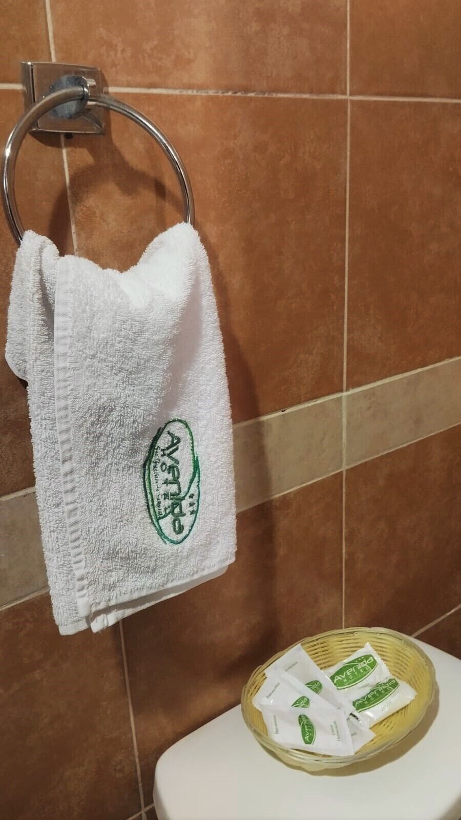 A white towel with a logo hanging on a metal ring, a basket with sachets on a white surface, and brown tiled walls.