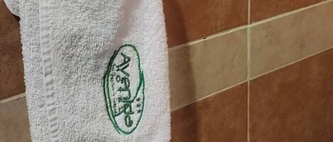 A white towel with a logo hanging on a metal ring, a basket with sachets on a white surface, and brown tiled walls.