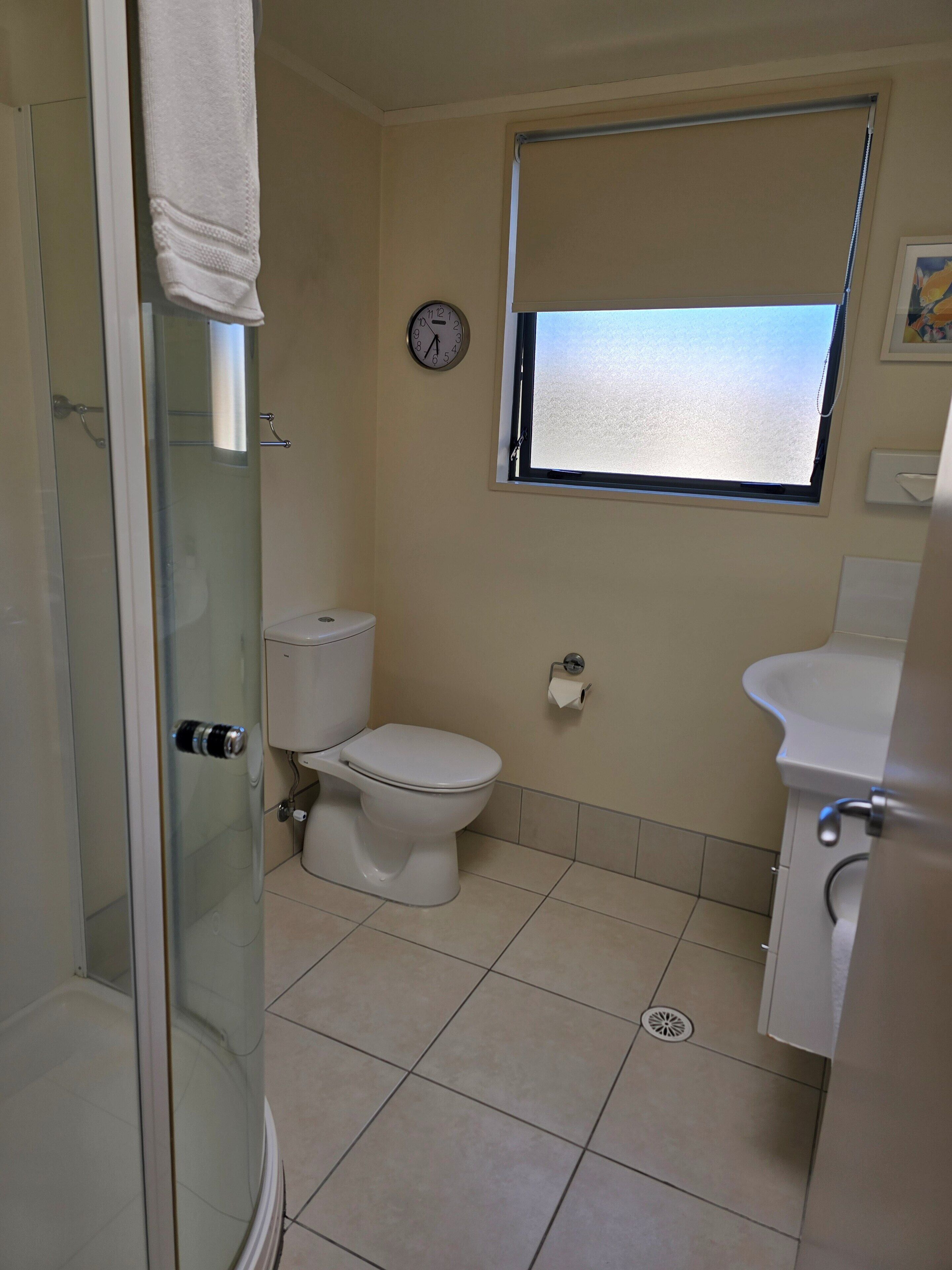 Un baño con inodoro, lavamanos, ventana con vistas, un reloj y zona de ducha.