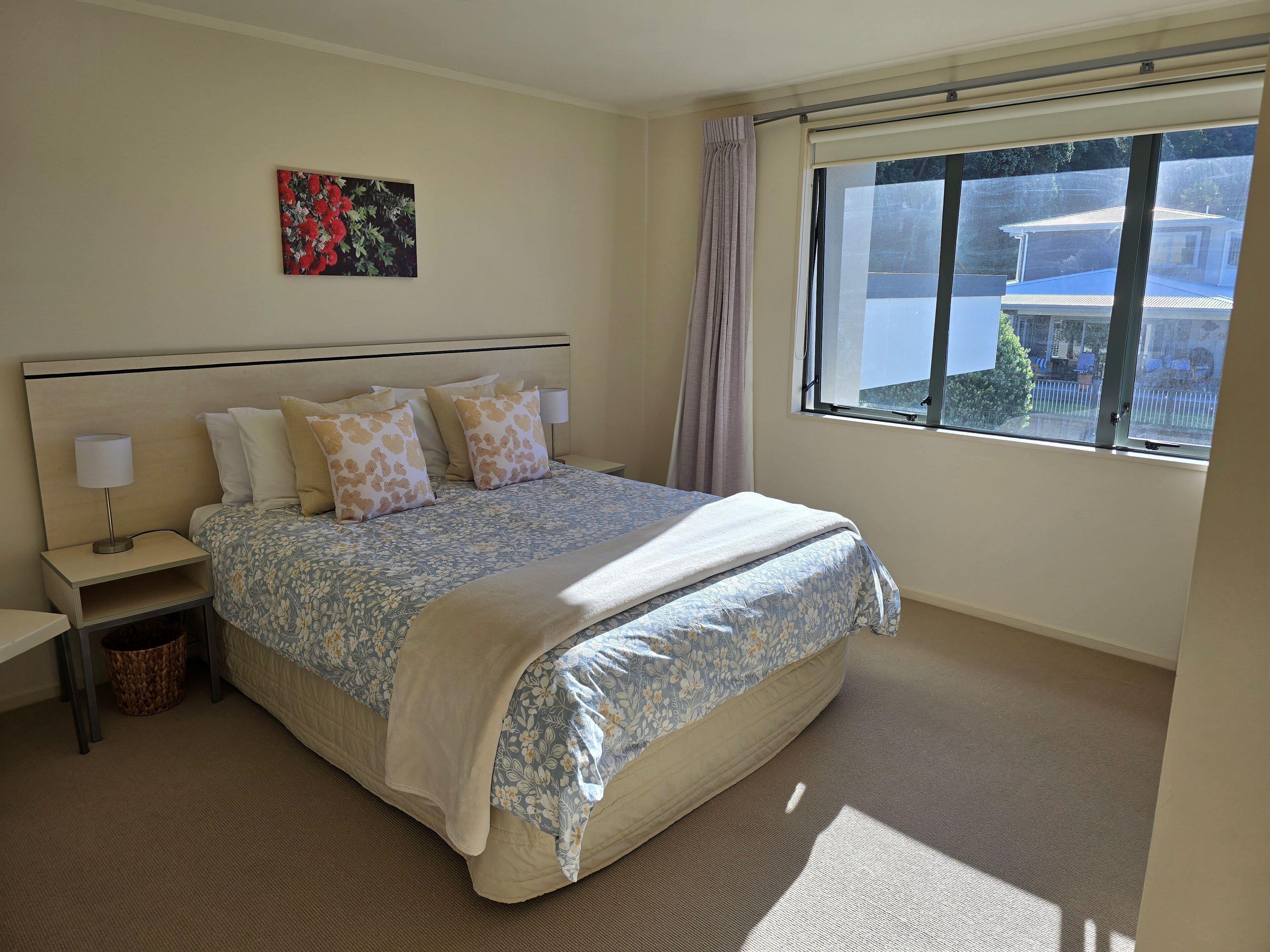 A neatly arranged bedroom with a large bed, a bedside table with a lamp, a small waste basket, and a window with a view of the outside.