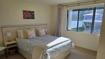 A neatly arranged bedroom with a large bed, a bedside table with a lamp, a small waste basket, and a window with a view of the outside.