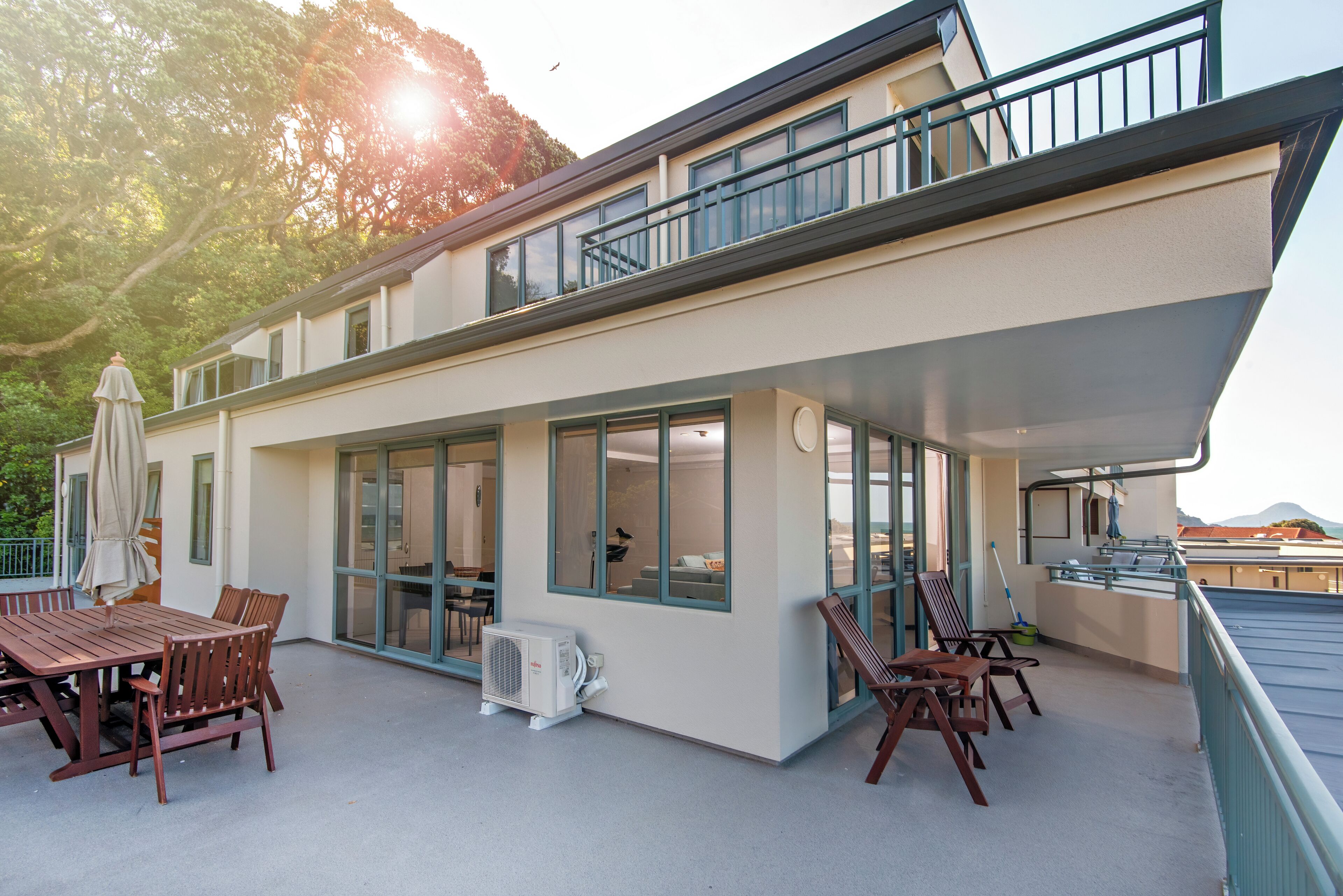 A modern building with a covered patio area featuring a wooden table and chairs, large windows, and a balcony with railings.