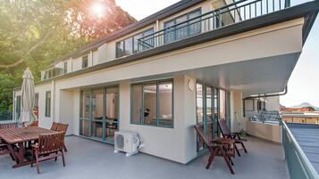 A modern building with a covered patio area featuring a wooden table and chairs, large windows, and a balcony with railings.