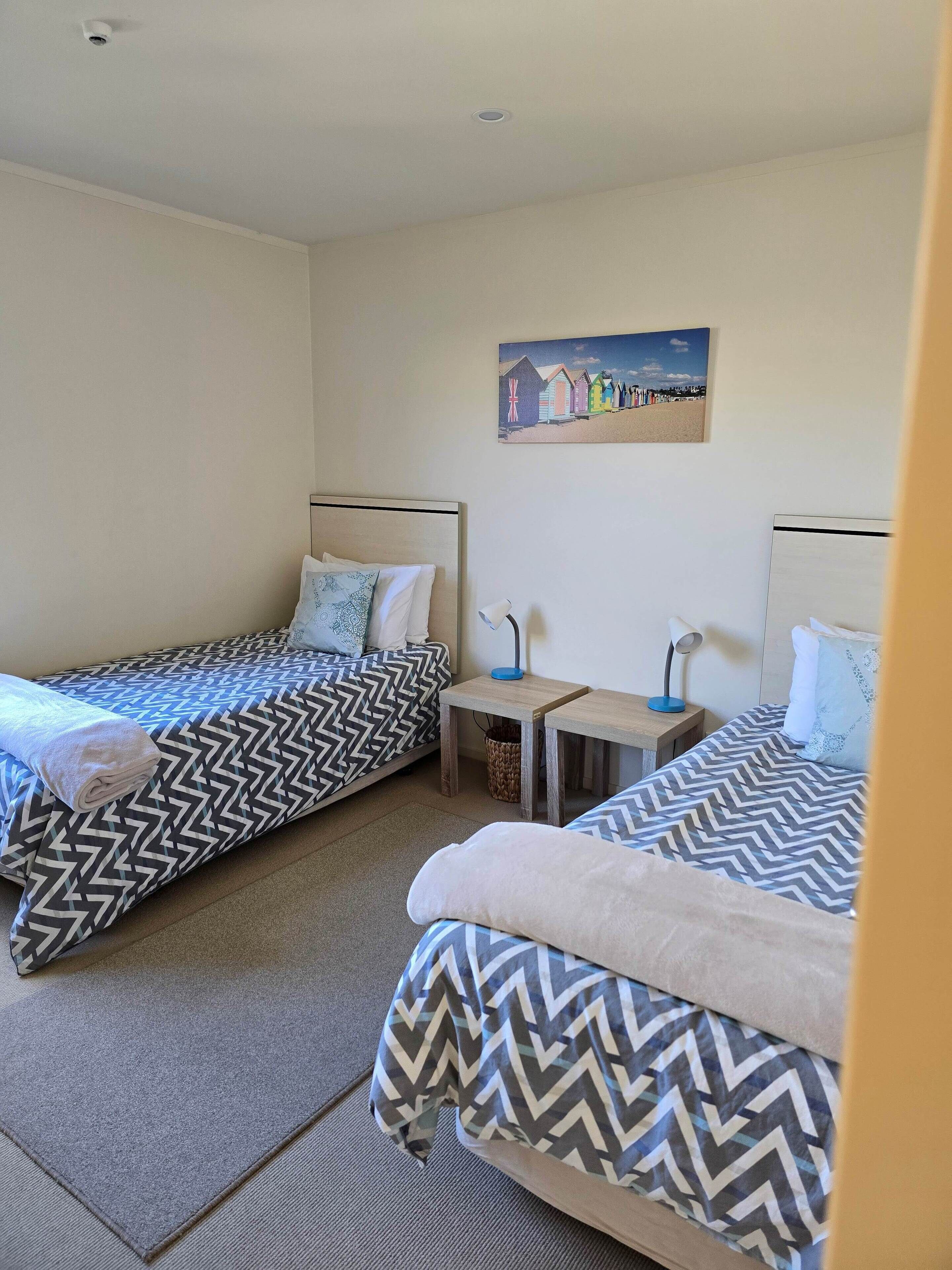 Een kamer met twee bedden, elk voorzien van een beddekleed met een patroon, een tafeltje met een lamp en een schilderij aan de muur.