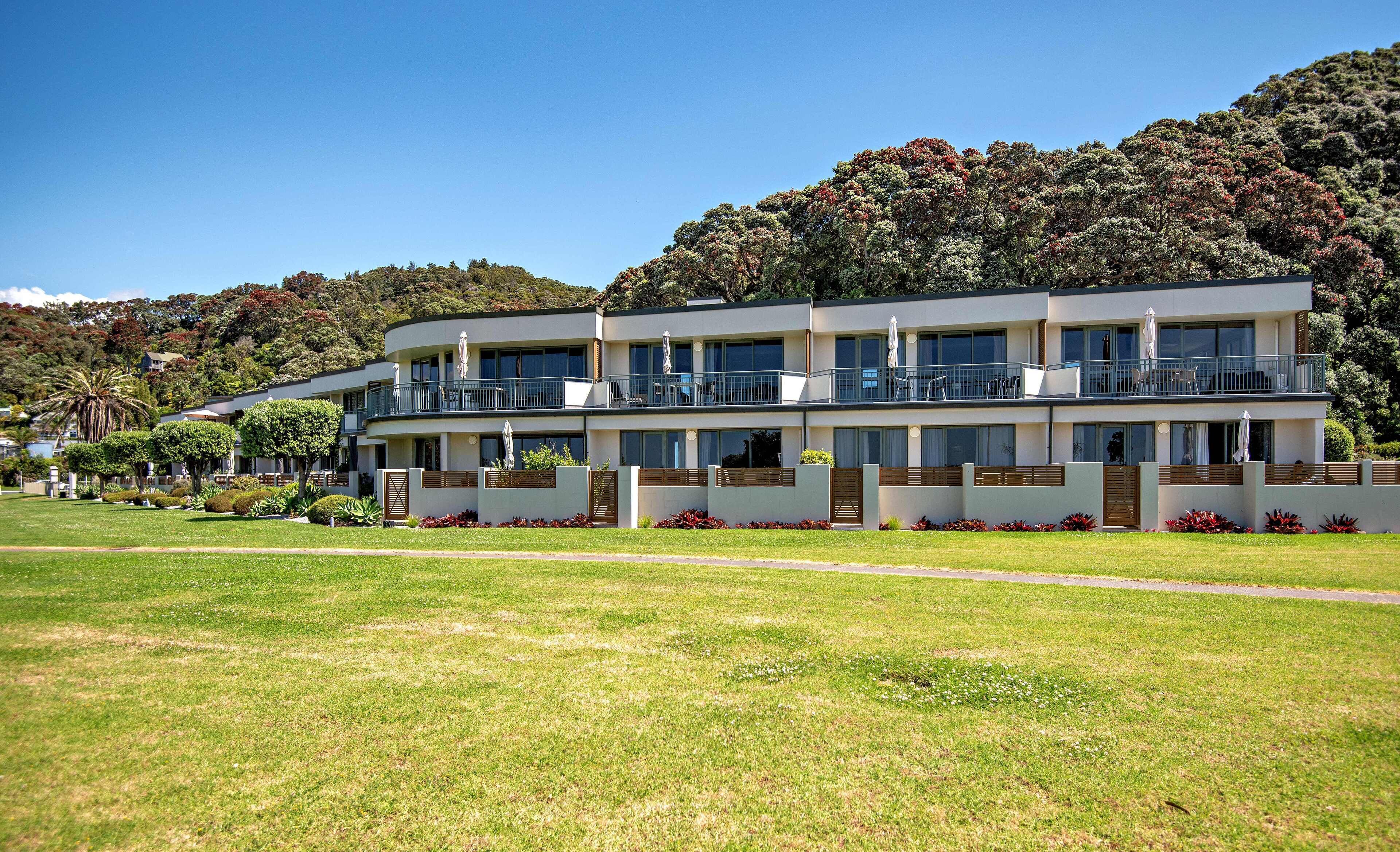 A modern two-story hotel with balconies, a well-maintained lawn, and a forested hillside in the background.