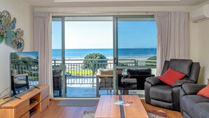 A modern living room with a balcony overlooking the ocean, featuring a leather sofa, a wooden coffee table, and a flat-screen TV. - Beachpoint Apartments (Ohope)