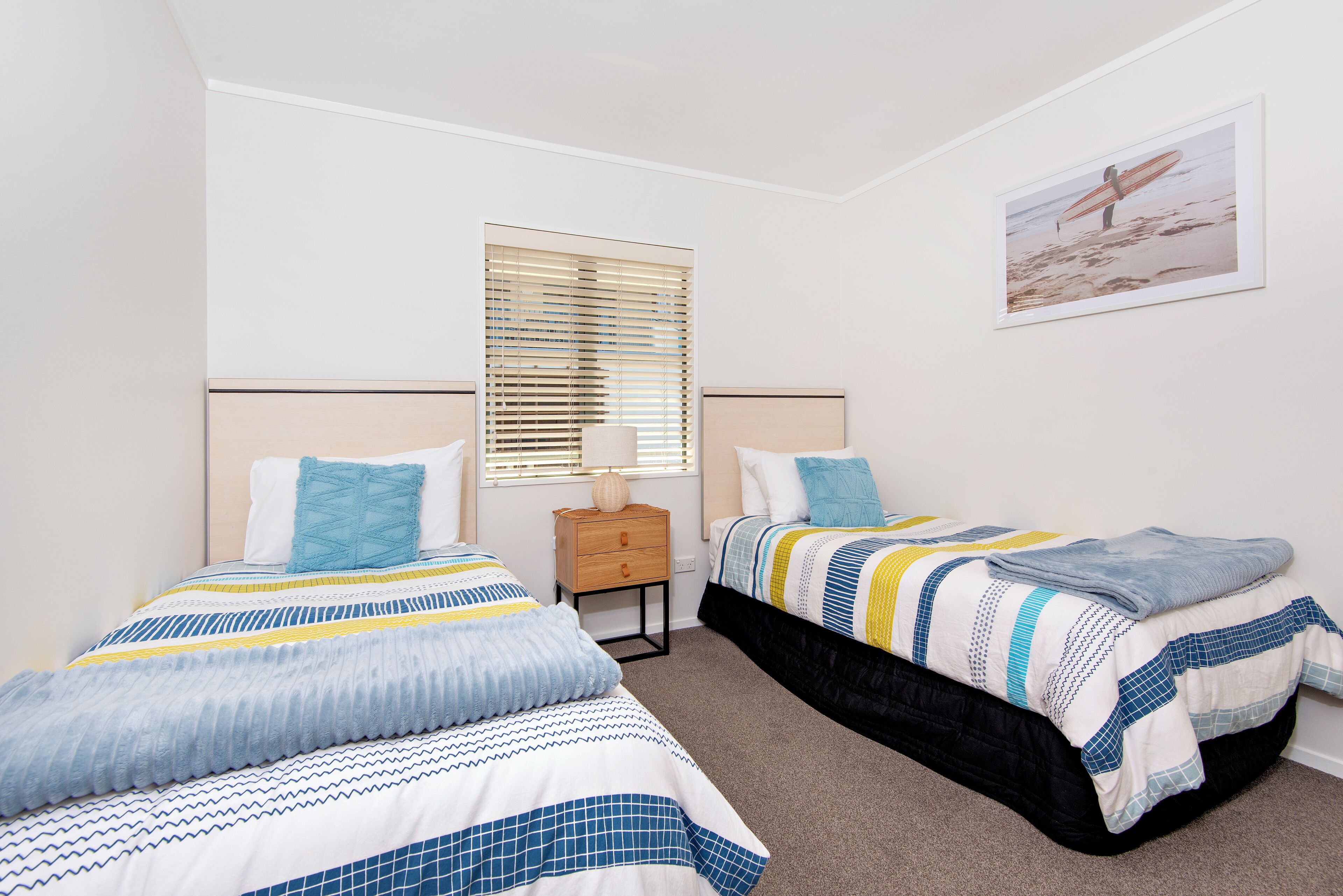 A bedroom with two beds, a nightstand, a window with blinds, and a framed picture on the wall.
