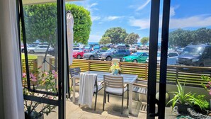 A view from a balcony with a table and chairs, a potted plant, and a view of a parking lot with various cars. - Beachpoint Apartments (Ohope)