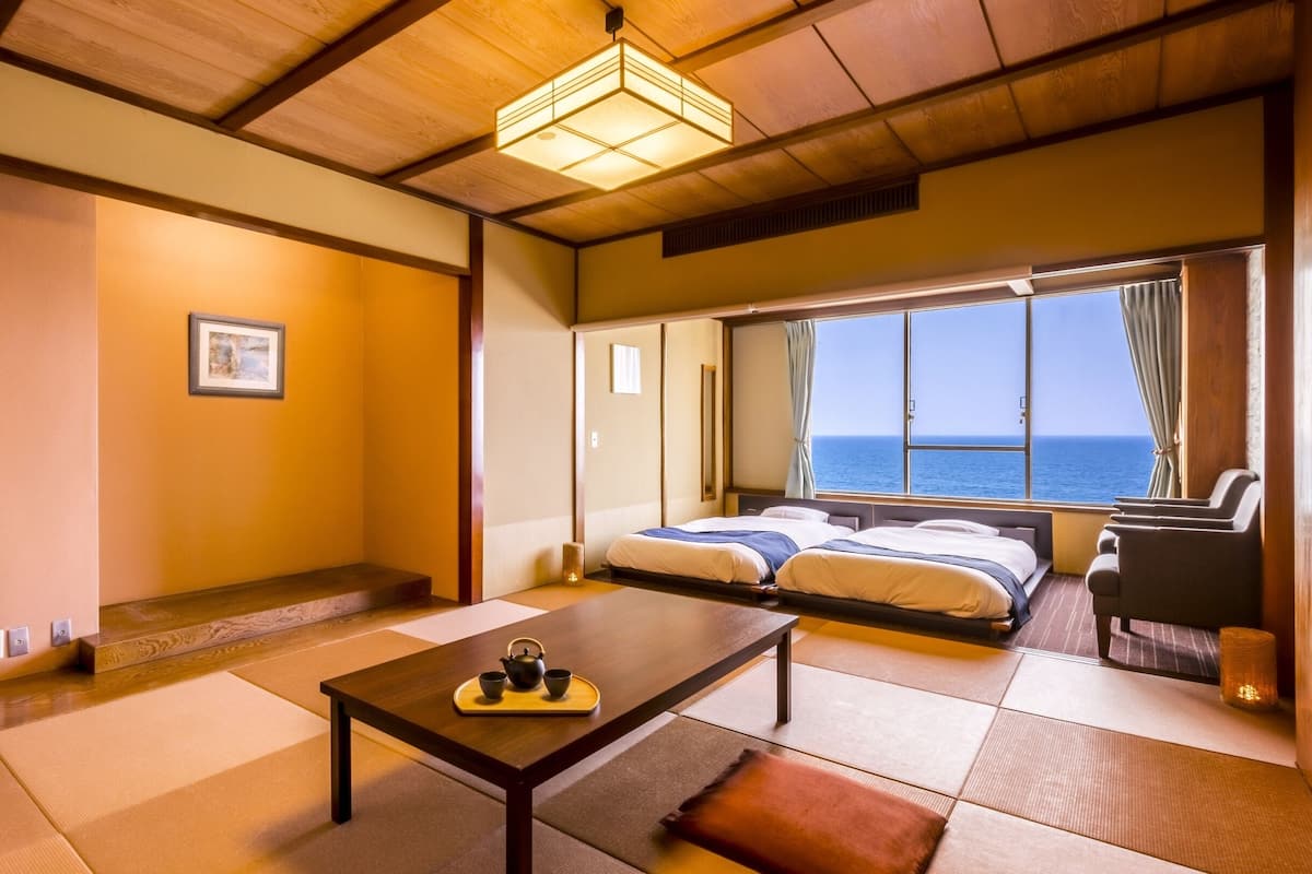A traditional Japanese-style room with tatami flooring, a low table with a tea set, two beds, and a view of the sea through large windows.