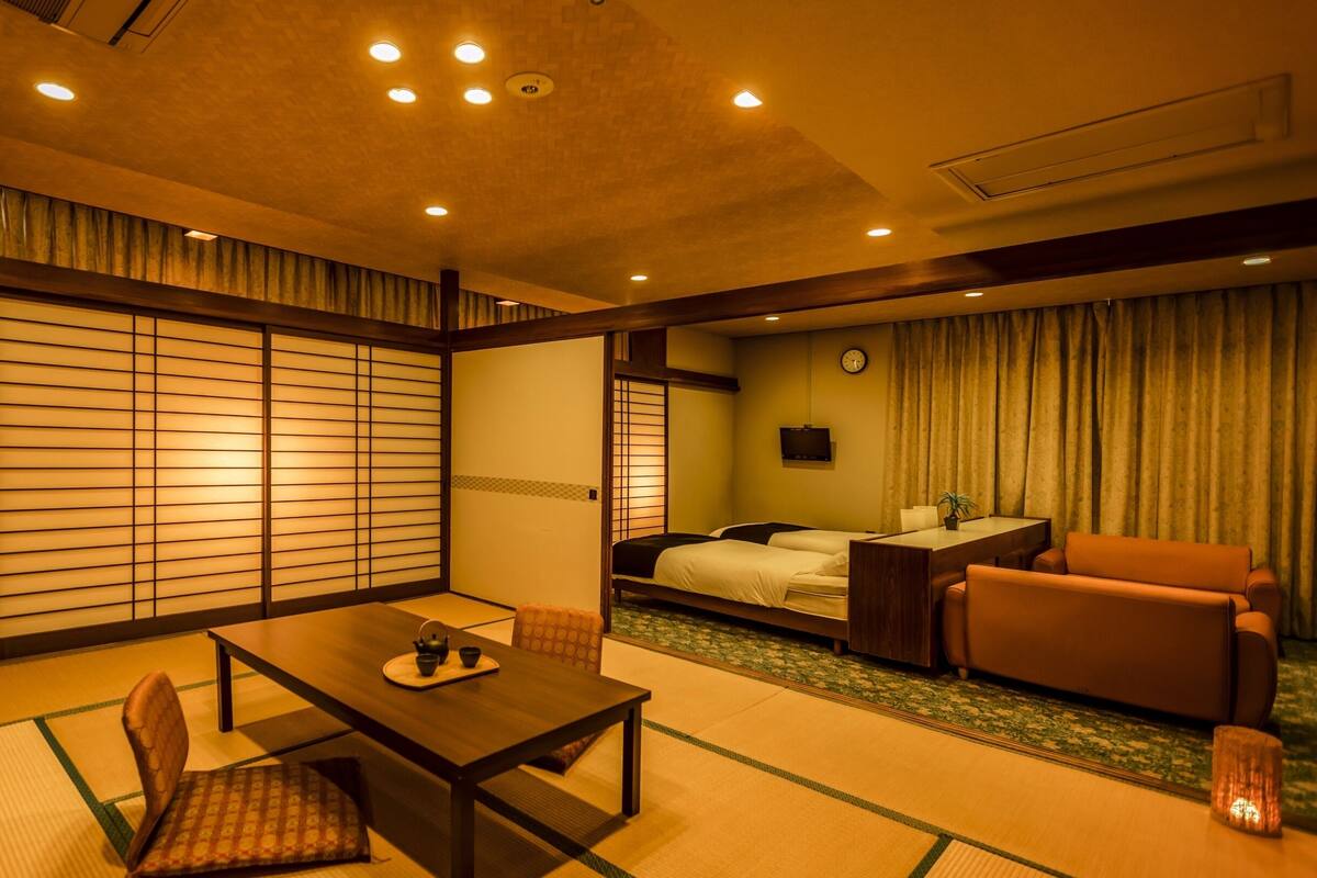 A traditional Japanese-style room with tatami flooring, a low table with a tea set, a bed with a wooden headboard, and a sofa.