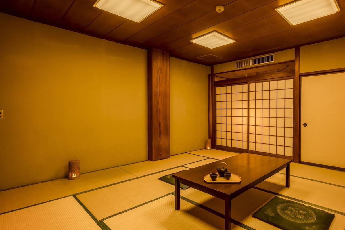 A traditional Japanese room with tatami flooring, a low wooden table with a tea set, sliding shoji doors, and a ceiling with recessed lighting.