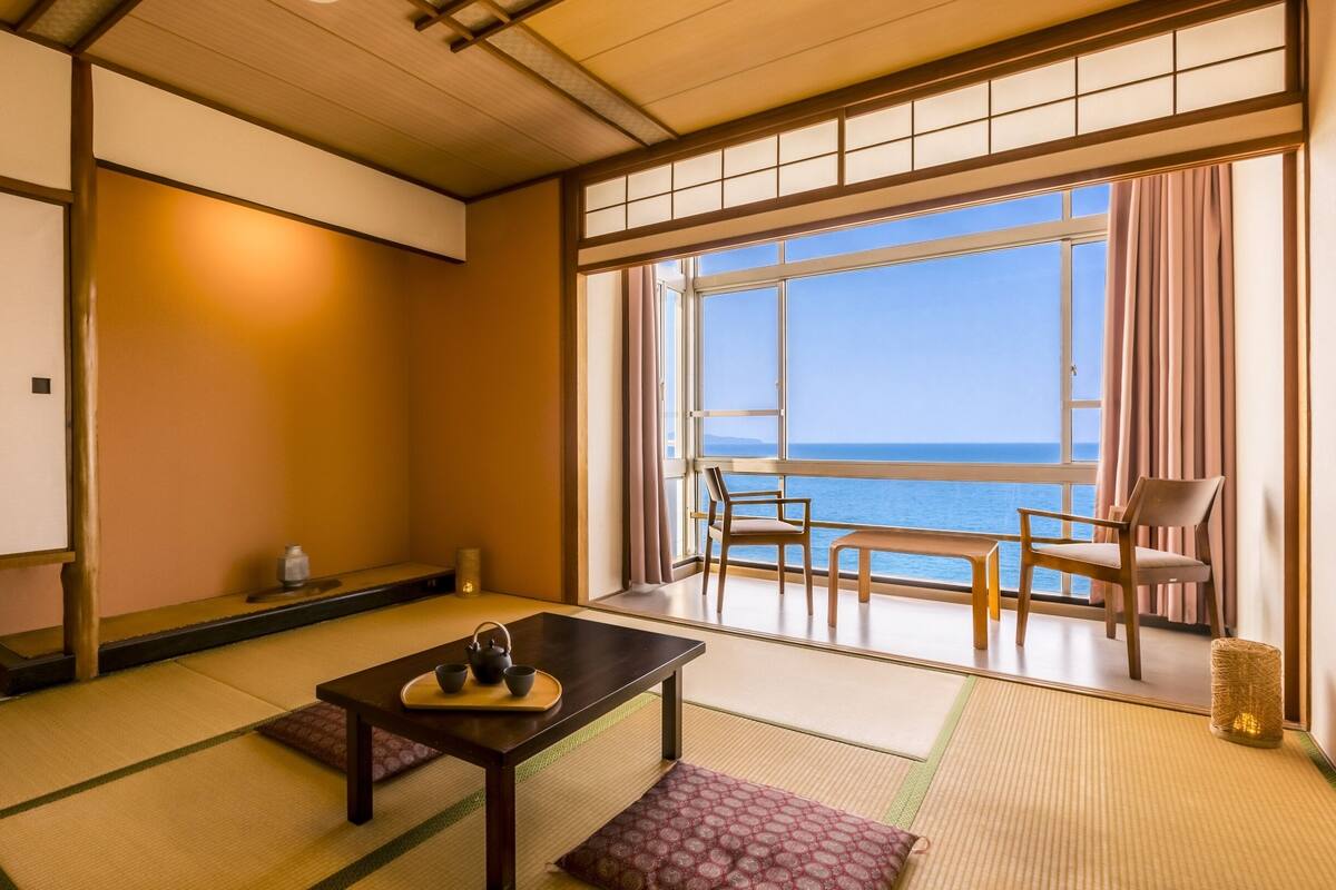 A traditional Japanese room with tatami flooring, a low table with tea set, and a view of the sea through large windows.