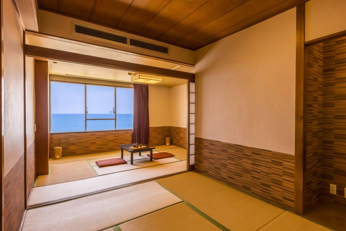 A traditional Japanese room with tatami flooring, a low table with a tea set, and a view of the sea through a large window.