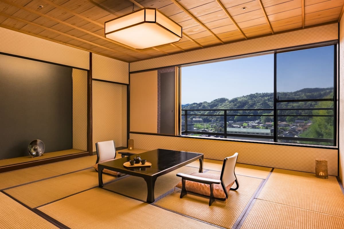 A traditional Japanese room with tatami flooring, a low table with a tea set, and a view of a town through large windows.