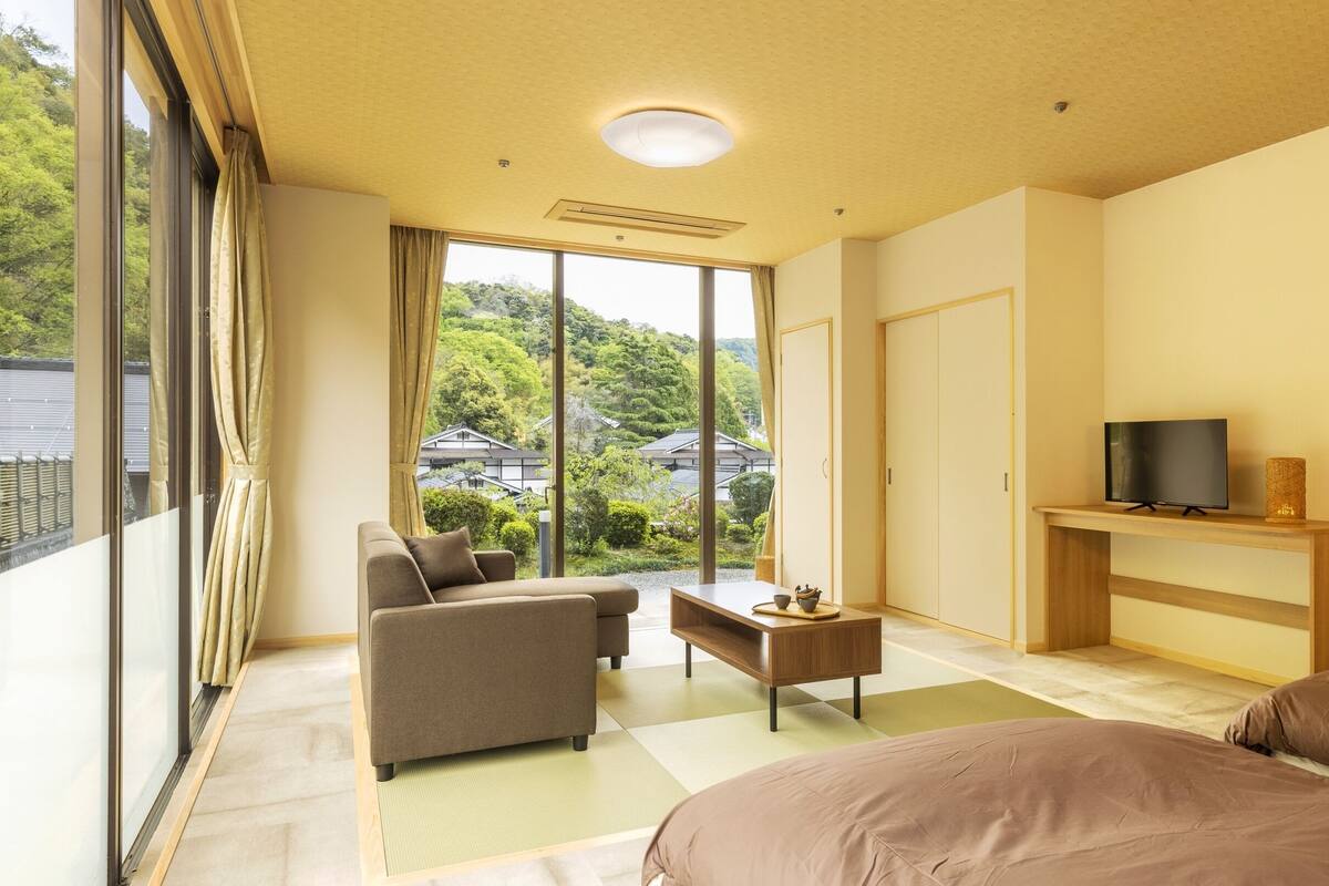 A modern hotel room with a sofa, a coffee table, a TV on a wooden stand, and a view of traditional buildings through large windows.