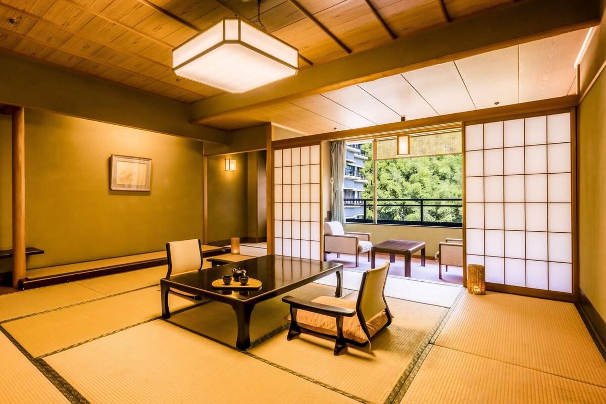 A traditional Japanese-style room with tatami flooring, a low table with tea set, and sliding doors leading to a balcony with a view of trees.