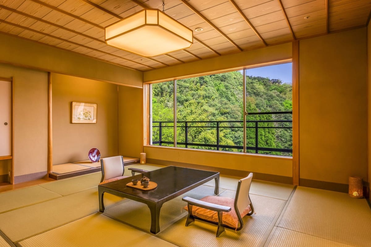 A traditional Japanese room with tatami flooring, a low table with a tray of tea utensils, a chair, a hanging light fixture, and a window with a view of a forest.