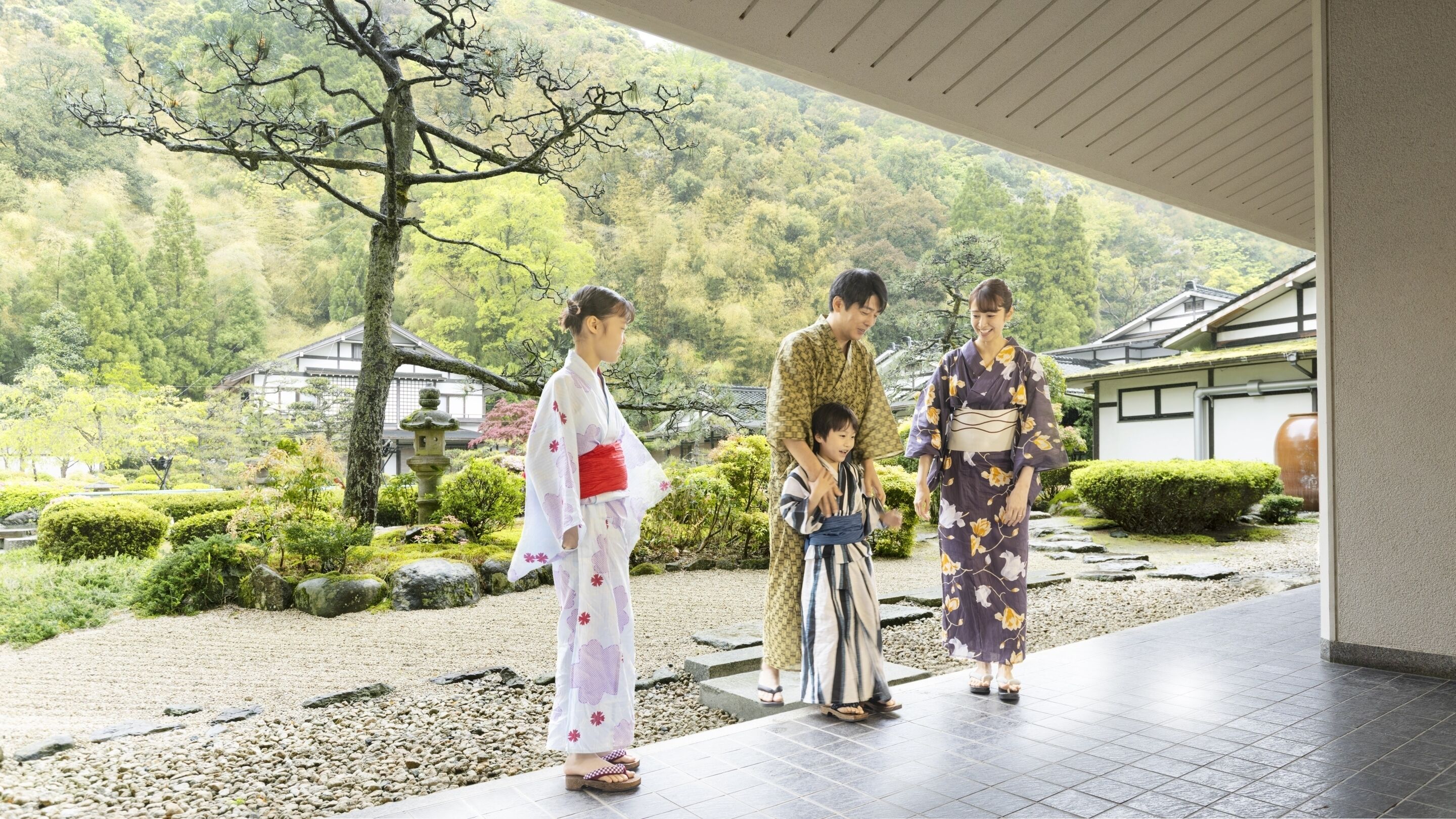 A traditional Japanese garden with a stone pathway, a wooden building, and a mountainous backdrop.
