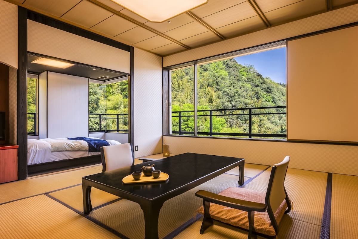 A traditional Japanese room with tatami flooring, a low table with a tea set, a bed with a view of a forest, and a sliding door.