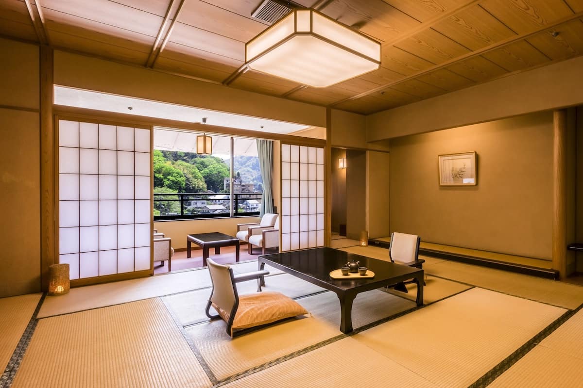 A traditional Japanese room with tatami flooring, a low table with tea set, sliding doors, and a view of a mountainous landscape.