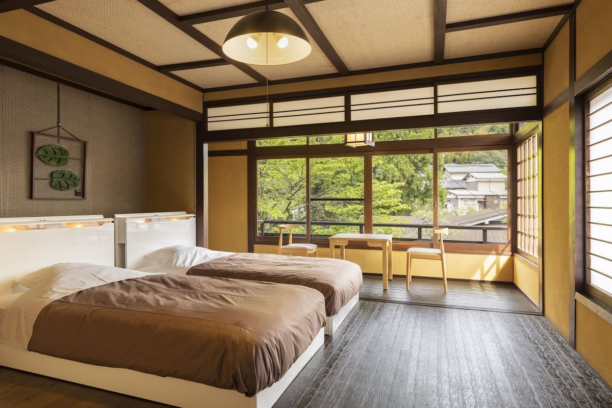 A traditional Japanese-style room with tatami flooring, a low wooden bed with two pillows, a small table with chairs, and a large window offering a view of greenery outside.