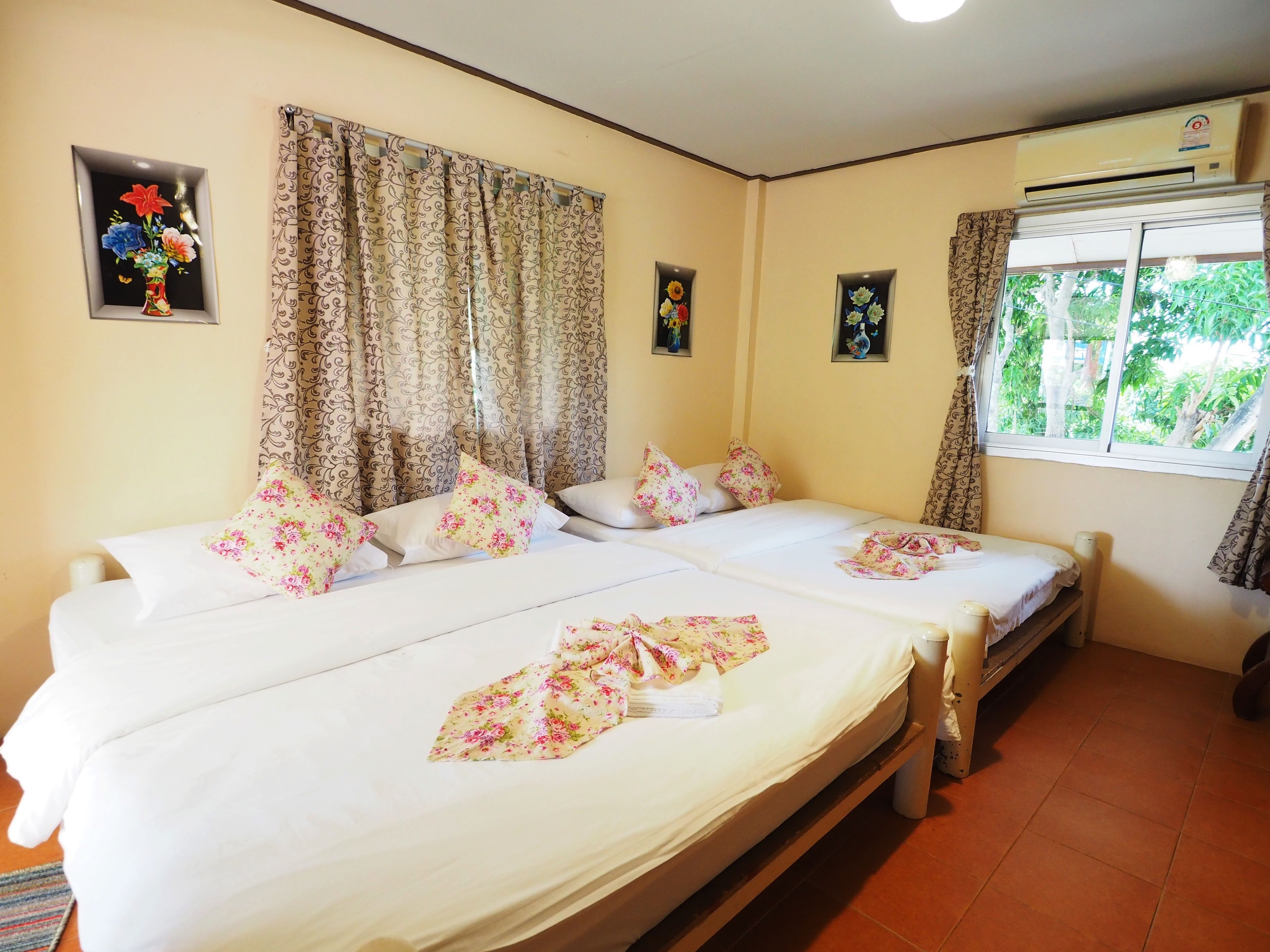 Una habitación con dos camas, ropa de cama estampada con motivos florales, una ventana con vistas a zonas verdes y un aire acondicionado instalado en la pared.