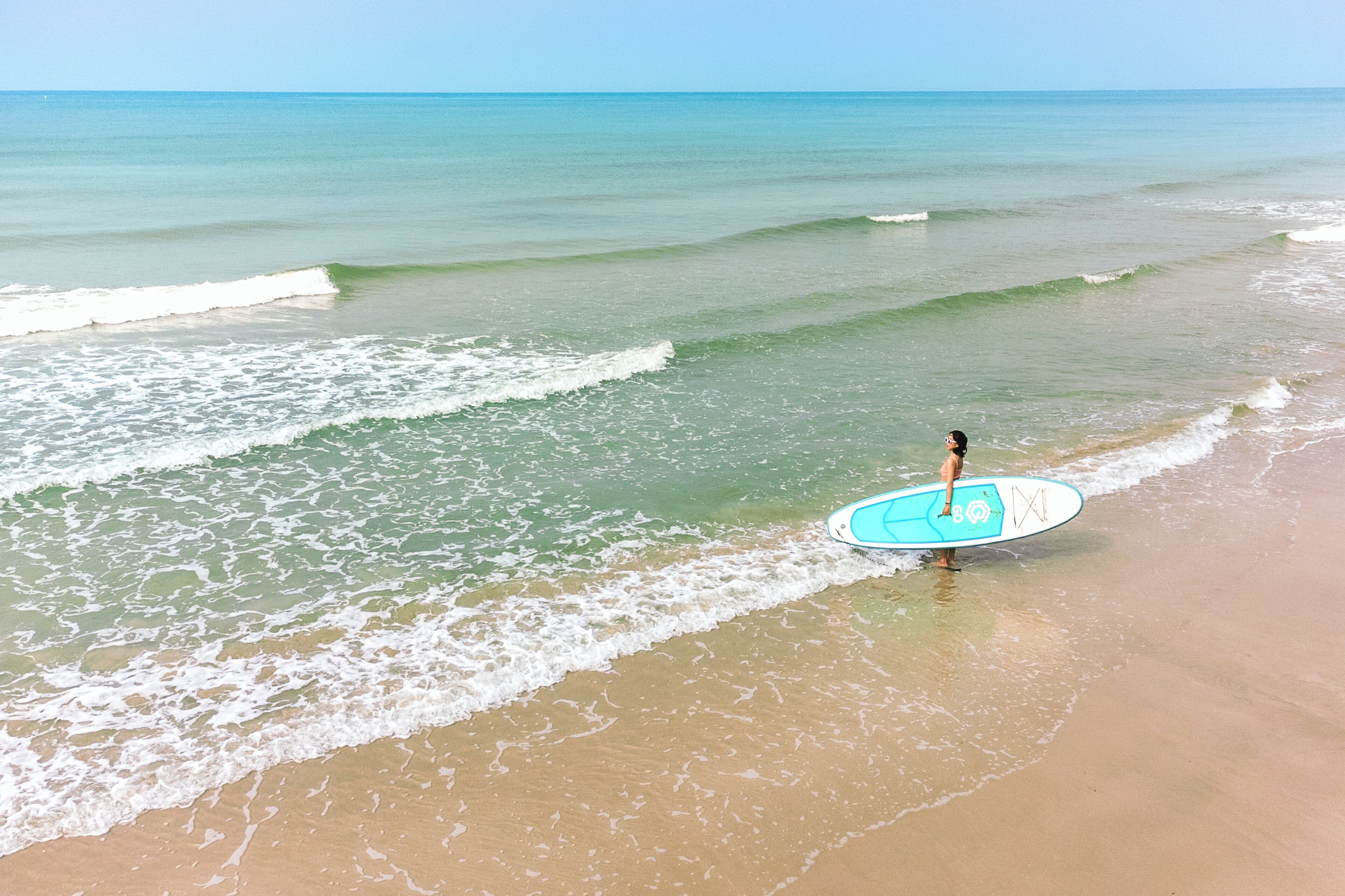 Una playa con agua cristalina y una persona sosteniendo una tabla de surf.