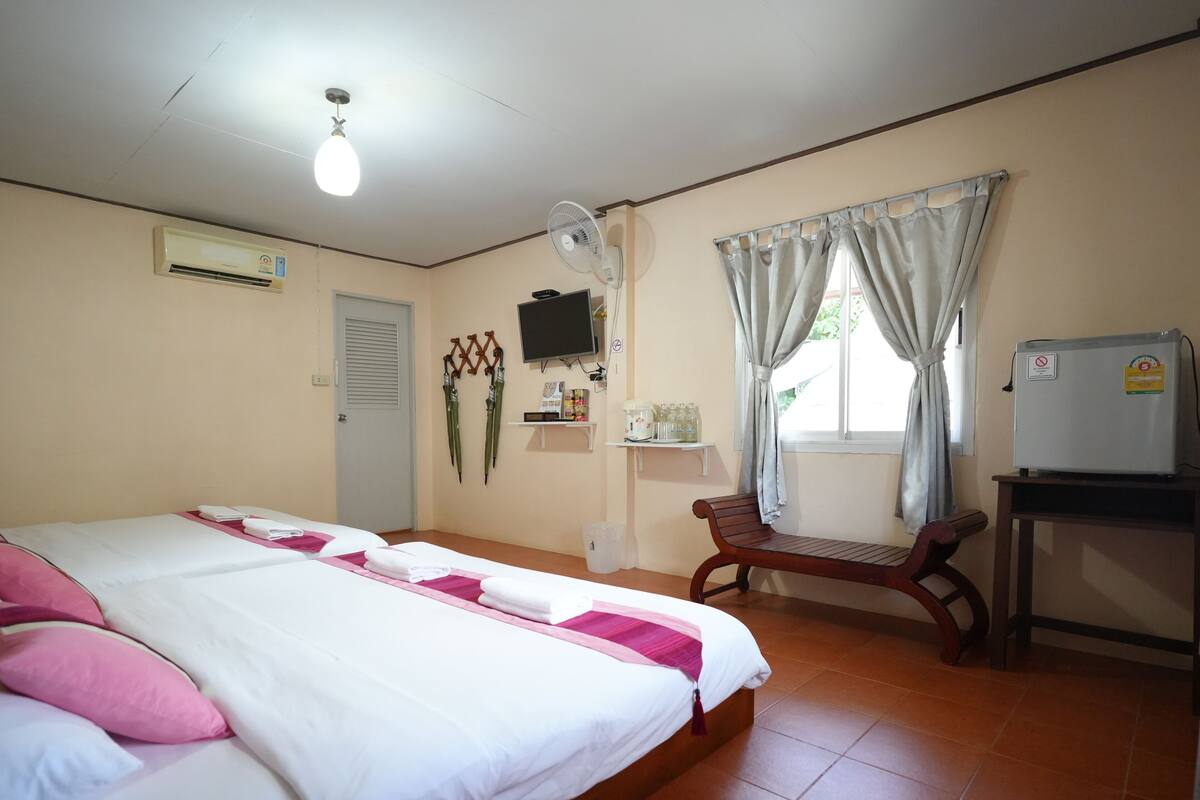 A hotel room with a bed, a desk, a chair, a TV, a fan, a window with curtains, and a wall-mounted air conditioner.
