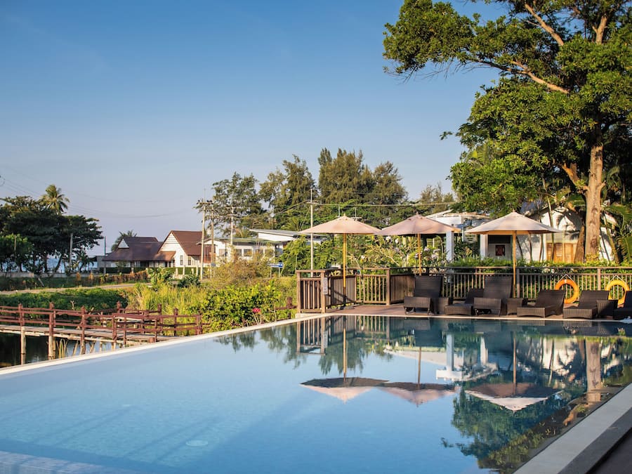 Piscina con sillas de descanso y sombrillas, rodeada de vegetación y una terraza de madera.