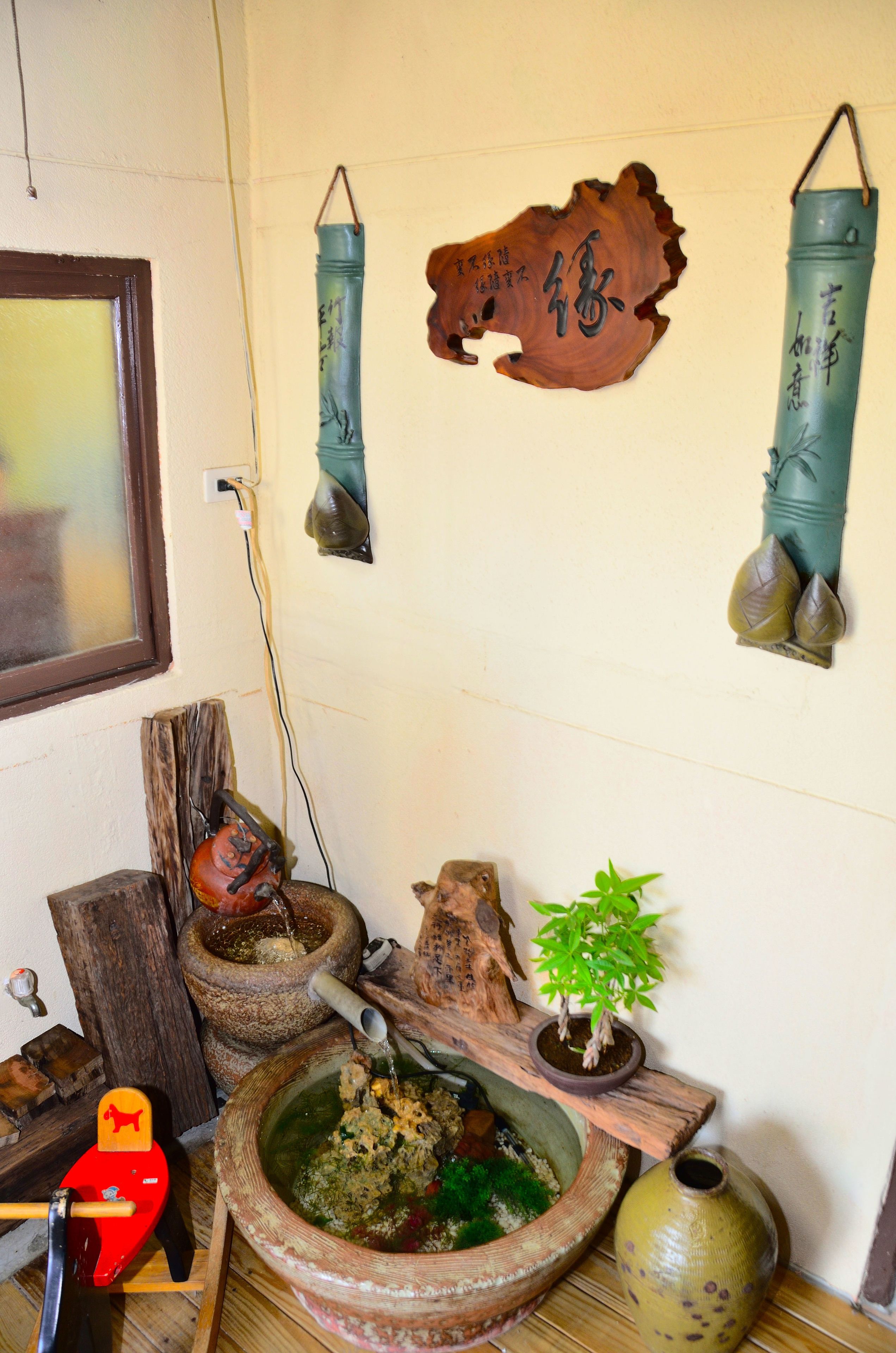 A traditional Japanese room with a wooden floor, a red rocking chair, a stone mortar and pestle, a wooden shelf with a bonsai tree, and a wall-mounted lantern.